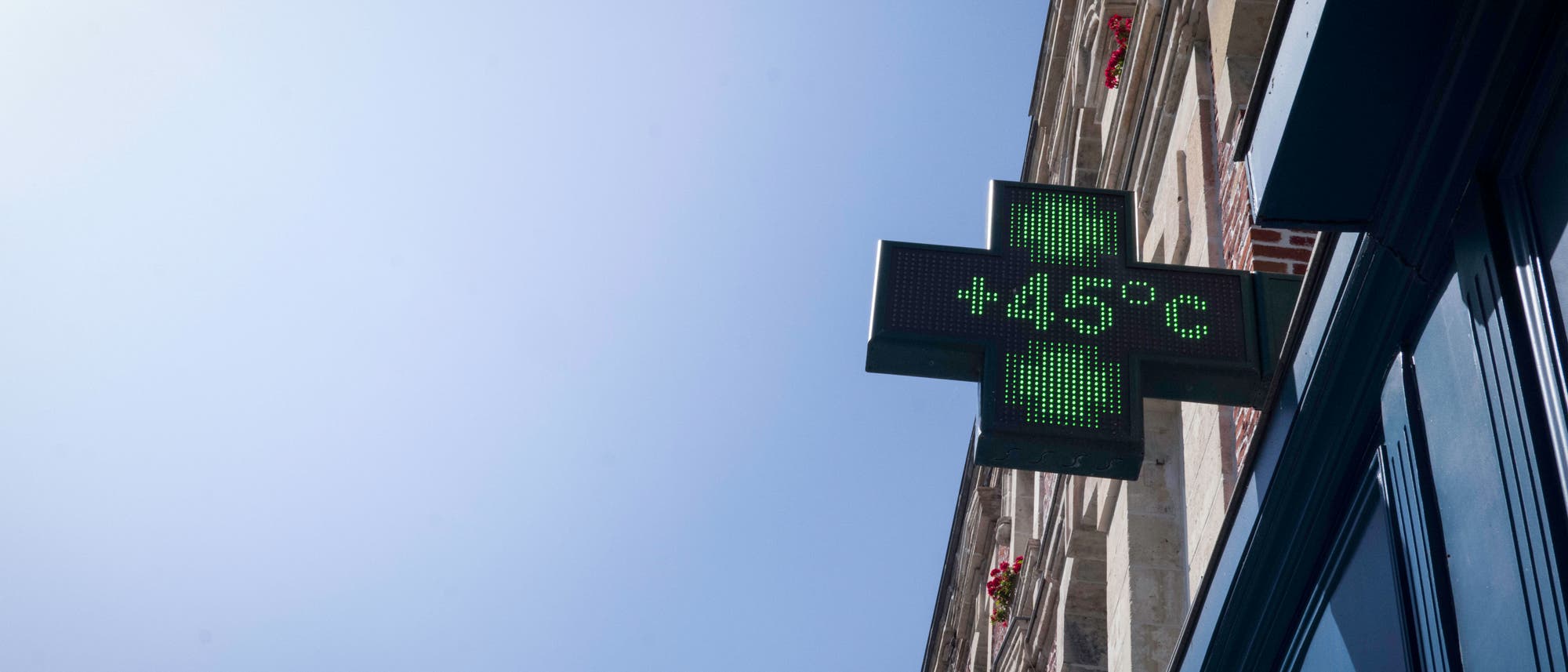 Ein digitales Schild in Form eines gr&uuml;nen Kreuzes zeigt die Temperatur von +45&deg;C an. Das Schild ist an der Fassade eines Geb&auml;udes angebracht, das in den Himmel ragt. Der Himmel ist klar und blau.