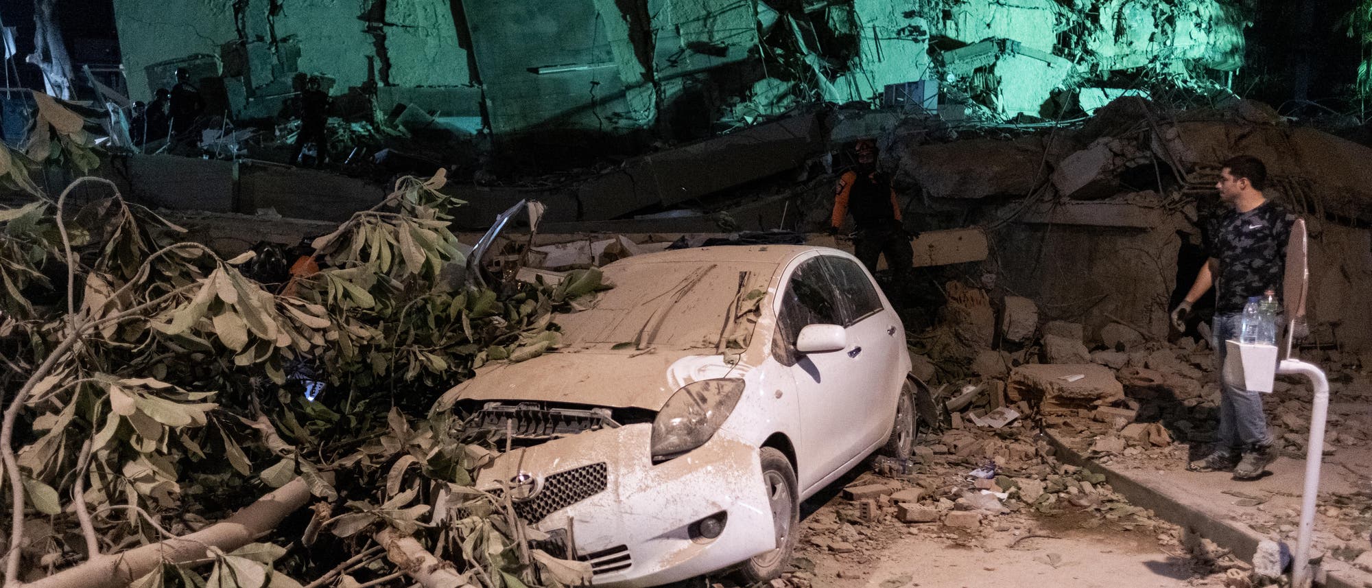 Ein zerst&ouml;rtes Geb&auml;ude liegt in Tr&uuml;mmern, umgeben von Schutt und umgest&uuml;rzten B&auml;umen. Ein wei&szlig;es Auto ist unter den Tr&uuml;mmern begraben. Eine Person steht rechts im Bild und betrachtet die Szene. Die Umgebung ist nachts beleuchtet, was die Zerst&ouml;rung hervorhebt.