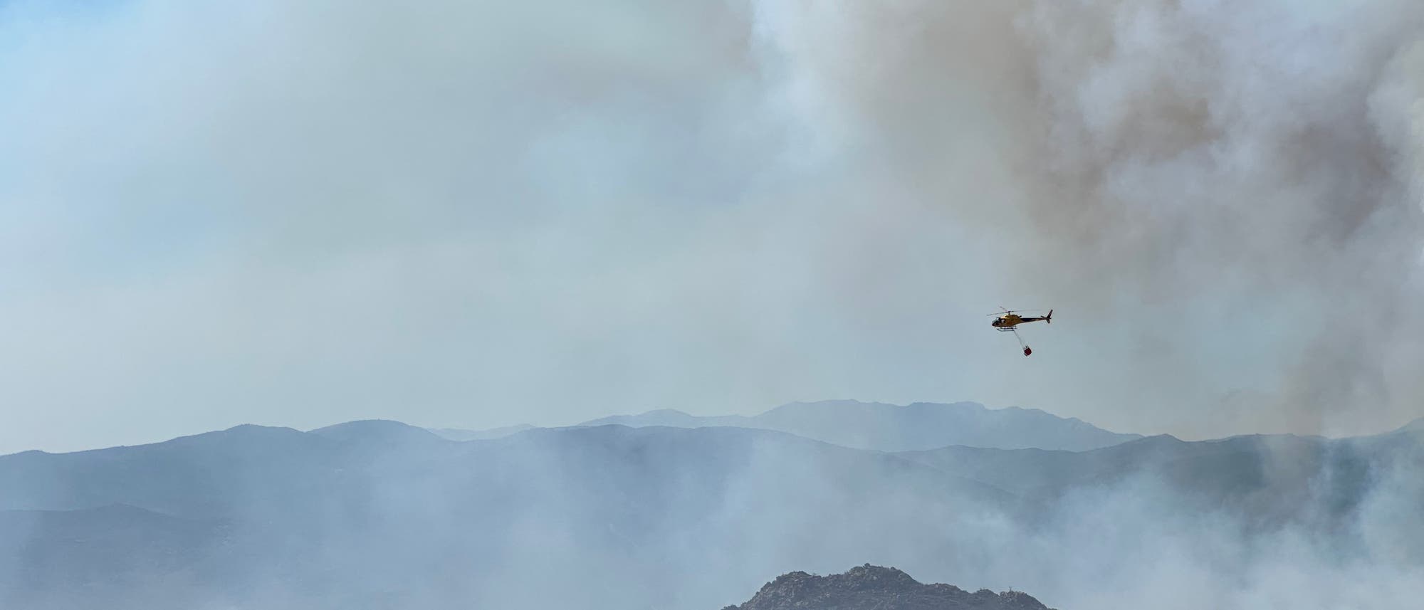 Ein Hubschrauber fliegt über eine Landschaft mit Hügeln und Bäumen, während er einen Wasserbehälter trägt. Im Hintergrund steigt Rauch in den Himmel, was auf einen Waldbrand hindeutet. Der Himmel ist blau, und die Szene zeigt die Bemühungen zur Brandbekämpfung in einer ländlichen Umgebung. Ein Hubschrauber fliegt über eine Landschaft mit Hügeln und Bäumen, während er einen Wasserbehälter trägt. Im Hintergrund steigt Rauch in den Himmel, was auf einen Waldbrand hindeutet. Der Himmel ist blau, und die Szene zeigt die Bemühungen zur Brandbekämpfung in einer ländlichen Umgebung.