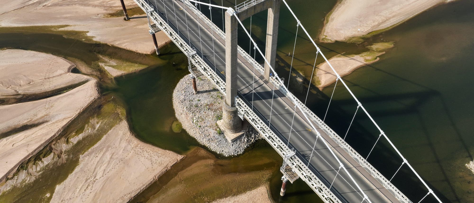 Luftaufnahme einer Hängebrücke, die über einen teilweise ausgetrockneten Fluss verläuft. Die Brücke ist aus Metall und hat zwei große Pfeiler, die sie stützen. Der Fluss zeigt sandige Ufer und flache Wasserstellen, die sich durch das Flussbett schlängeln. Die Umgebung wirkt trocken und karg. Luftaufnahme einer Hängebrücke, die über einen teilweise ausgetrockneten Fluss verläuft. Die Brücke ist aus Metall und hat zwei große Pfeiler, die sie stützen. Der Fluss zeigt sandige Ufer und flache Wasserstellen, die sich durch das Flussbett schlängeln. Die Umgebung wirkt trocken und karg.