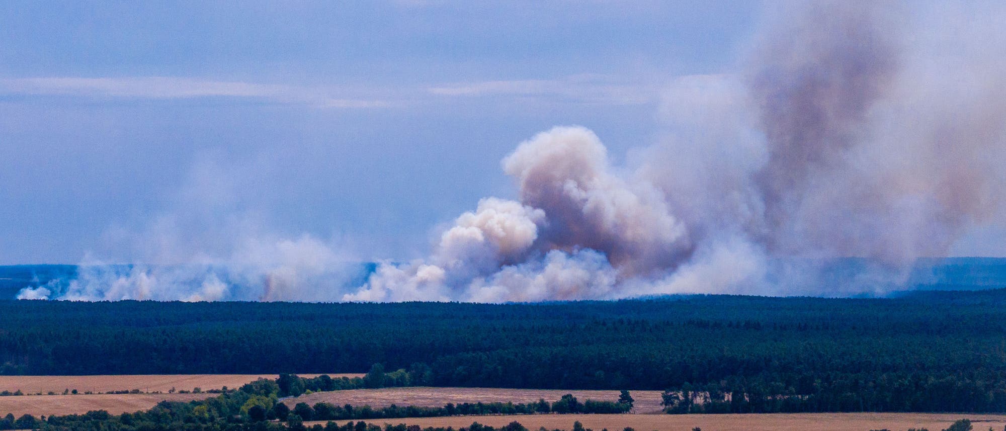 Landschaftsfoto mit einem Waldbrand im Hintergrund. Dichte Rauchwolken steigen aus einem Waldgebiet auf, während im Vordergrund Felder und eine kleine Ansammlung von Häusern zu sehen sind. Der Himmel ist leicht bewölkt. Landschaftsfoto mit einem Waldbrand im Hintergrund. Dichte Rauchwolken steigen aus einem Waldgebiet auf, während im Vordergrund Felder und eine kleine Ansammlung von Häusern zu sehen sind. Der Himmel ist leicht bewölkt.