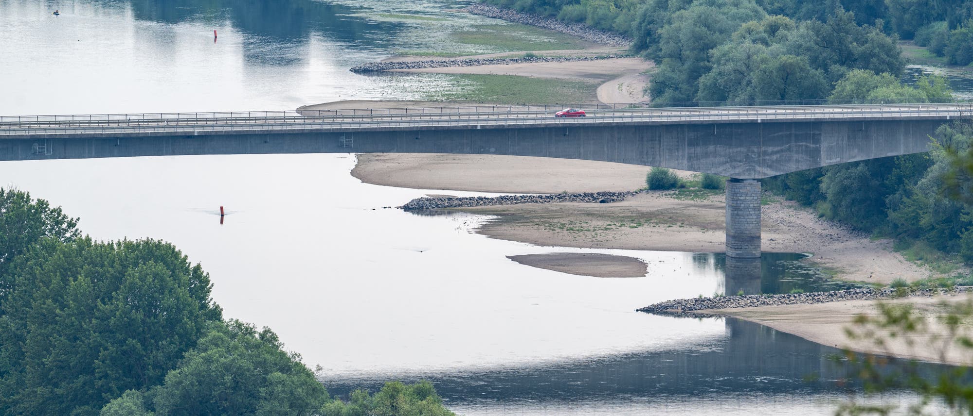 Eine Br&uuml;cke &uuml;berspannt einen ruhigen Fluss, umgeben von &uuml;ppigem Gr&uuml;n und B&auml;umen. Auf der Br&uuml;cke f&auml;hrt ein rotes Auto. Der Fluss hat niedrigen Wasserstand, wodurch Sandb&auml;nke sichtbar werden. Im Hintergrund sind dichte W&auml;lder und landwirtschaftliche Fl&auml;chen zu sehen.