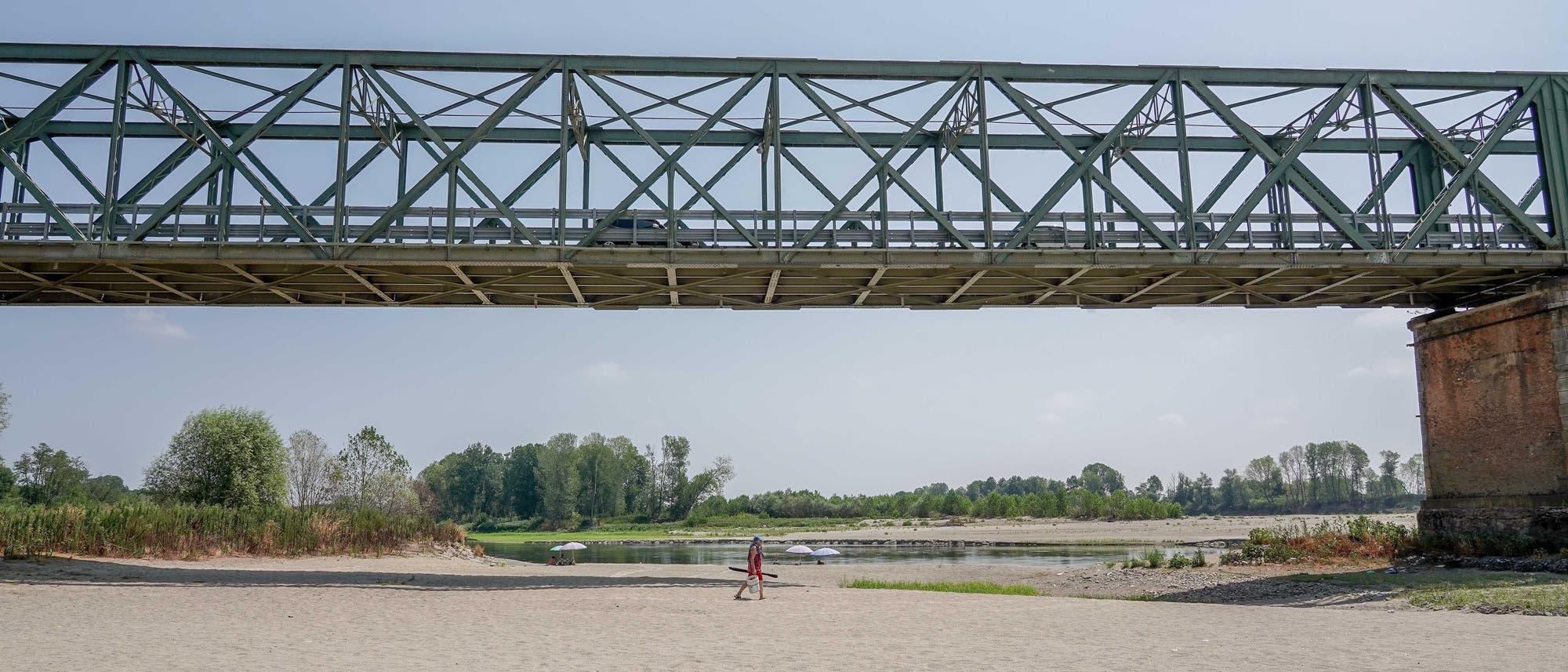 Eine Metallbrücke erstreckt sich über einen breiten, sandigen Flussstrand. Unter der Brücke geht eine Person in roter Kleidung. Im Hintergrund sind Bäume und ein ruhiger Fluss zu sehen. Der Himmel ist klar und blau. Eine Metallbrücke erstreckt sich über einen breiten, sandigen Flussstrand. Unter der Brücke geht eine Person in roter Kleidung. Im Hintergrund sind Bäume und ein ruhiger Fluss zu sehen. Der Himmel ist klar und blau.