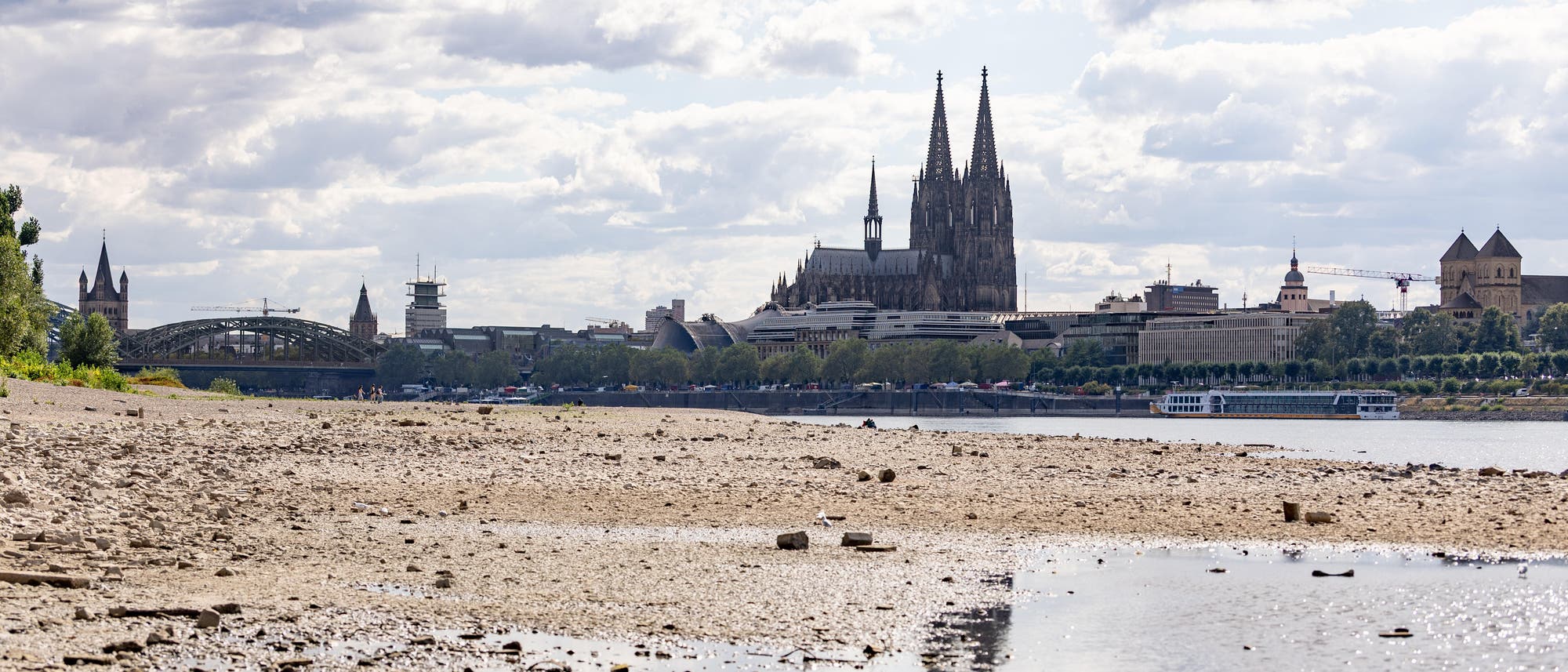 Blick auf eine Stadtlandschaft mit einem Fluss im Vordergrund und einer markanten gotischen Kathedrale im Hintergrund. Der Himmel ist bewölkt, und das Ufer ist steinig und trocken. Eine Brücke und mehrere historische Gebäude sind ebenfalls sichtbar. Ein Boot fährt auf dem Fluss. Blick auf eine Stadtlandschaft mit einem Fluss im Vordergrund und einer markanten gotischen Kathedrale im Hintergrund. Der Himmel ist bewölkt, und das Ufer ist steinig und trocken. Eine Brücke und mehrere historische Gebäude sind ebenfalls sichtbar. Ein Boot fährt auf dem Fluss.