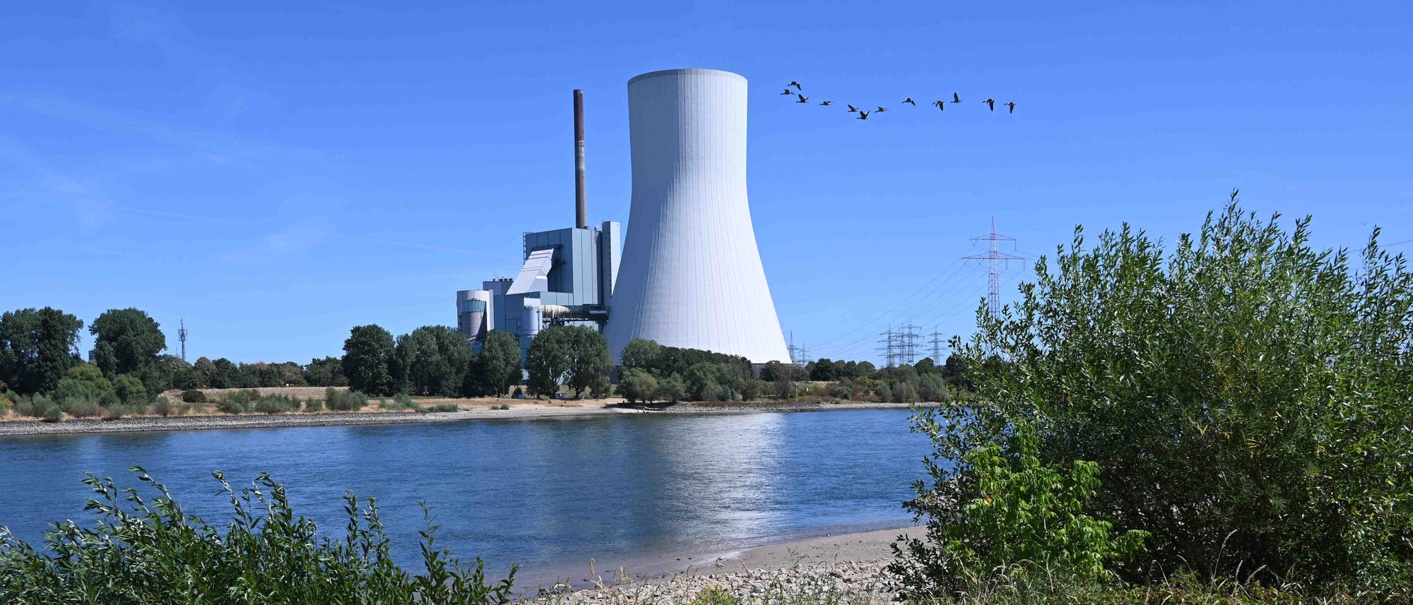 Ein großes Kraftwerk mit einem markanten Kühlturm steht am Ufer eines Flusses. Im Vordergrund sind Büsche und Bäume zu sehen, während eine Gruppe von Vögeln über den klaren blauen Himmel fliegt. Stromleitungen verlaufen in der Ferne, und das Wasser des Flusses spiegelt die Umgebung wider. Ein großes Kraftwerk mit einem markanten Kühlturm steht am Ufer eines Flusses. Im Vordergrund sind Büsche und Bäume zu sehen, während eine Gruppe von Vögeln über den klaren blauen Himmel fliegt. Stromleitungen verlaufen in der Ferne, und das Wasser des Flusses spiegelt die Umgebung wider.