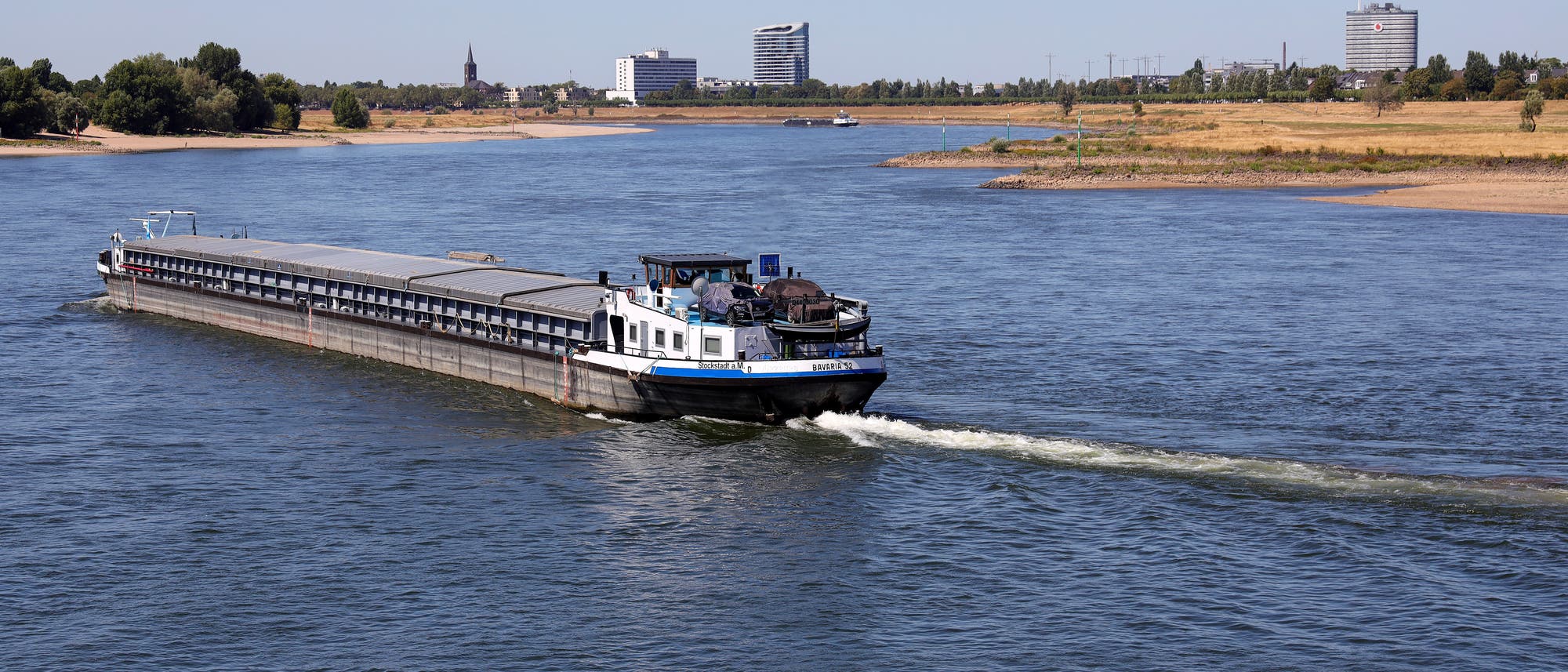 Ein Frachtschiff fährt auf einem breiten Fluss, umgeben von einer Landschaft mit Bäumen und modernen Gebäuden im Hintergrund. Der Himmel ist klar und blau. Das Schiff zieht eine leichte Welle hinter sich her. Ein Frachtschiff fährt auf einem breiten Fluss, umgeben von einer Landschaft mit Bäumen und modernen Gebäuden im Hintergrund. Der Himmel ist klar und blau. Das Schiff zieht eine leichte Welle hinter sich her.