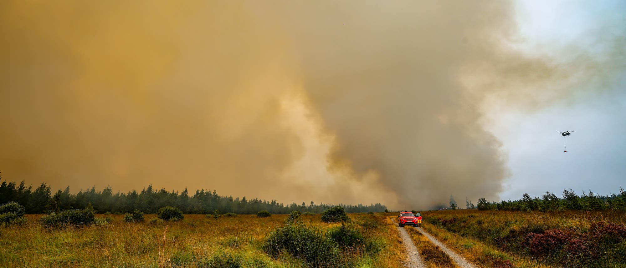 Eine gro&szlig;e Rauchwolke steigt &uuml;ber dem Hohen Venn auf, w&auml;hrend ein rotes Einsatzfahrzeug auf einem Schotterweg in Richtung des Rauchs f&auml;hrt. Ein Hubschrauber fliegt am Himmel und tr&auml;gt einen Wasserbeh&auml;lter, um den Brand zu bek&auml;mpfen. Die Szene zeigt eine Landschaft mit Gras und B&auml;umen unter einem bew&ouml;lkten Himmel.