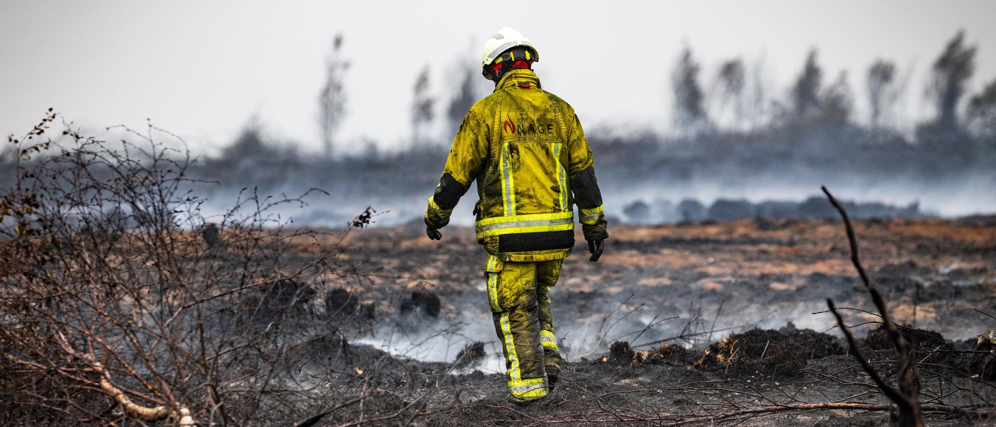 Ein Feuerwehrmann in gelber Schutzkleidung und Helm geht über ein verbranntes, rauchendes Feld. Im Hintergrund sind verkohlte Bäume und ein grauer Himmel zu sehen. Die Szene vermittelt den Eindruck eines kürzlich gelöschten Brandes. Ein Feuerwehrmann in gelber Schutzkleidung und Helm geht über ein verbranntes, rauchendes Feld. Im Hintergrund sind verkohlte Bäume und ein grauer Himmel zu sehen. Die Szene vermittelt den Eindruck eines kürzlich gelöschten Brandes.