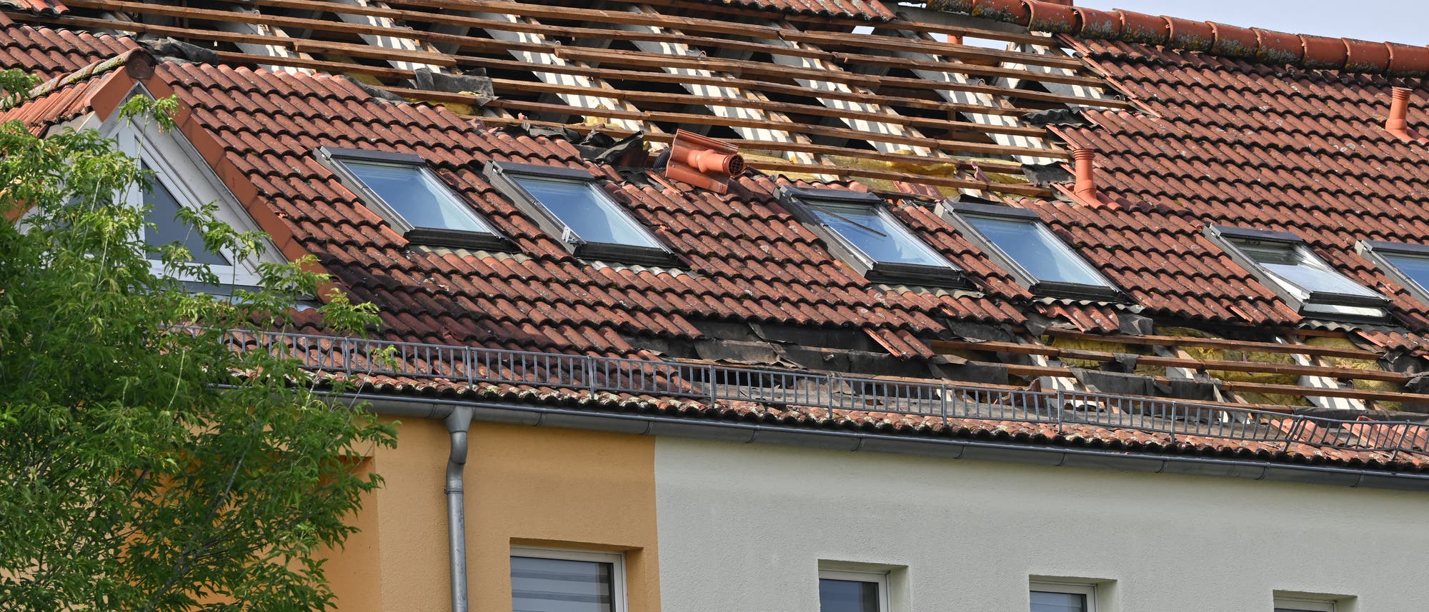 Ein Wohngebäude mit einem beschädigten Ziegeldach, bei dem mehrere Ziegel fehlen und die Dachkonstruktion sichtbar ist. Einige Dachfenster sind intakt, während andere von den Schäden betroffen sind. Im Vordergrund ist ein Baum zu sehen, der teilweise das Gebäude verdeckt. Das Bild zeigt die Auswirkungen von Sturmschäden auf die Dachstruktur. Ein Wohngebäude mit einem beschädigten Ziegeldach, bei dem mehrere Ziegel fehlen und die Dachkonstruktion sichtbar ist. Einige Dachfenster sind intakt, während andere von den Schäden betroffen sind. Im Vordergrund ist ein Baum zu sehen, der teilweise das Gebäude verdeckt. Das Bild zeigt die Auswirkungen von Sturmschäden auf die Dachstruktur.