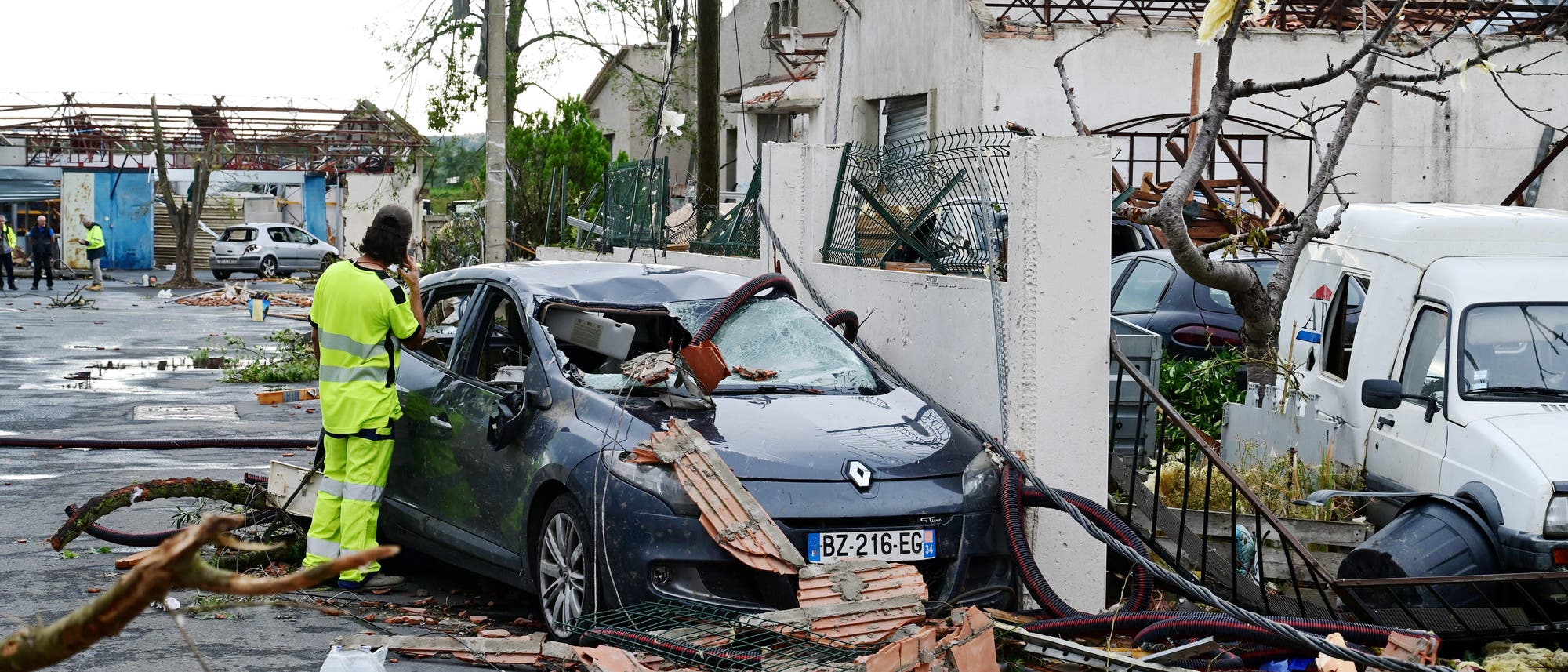 Eine Person in gelber Warnkleidung fotografiert ein beschädigtes Auto, das von Trümmern bedeckt ist. Im Hintergrund sind zerstörte Gebäude und umgestürzte Bäume zu sehen. Die Szene zeigt die Auswirkungen eines schweren Sturms oder Tornados. Auf dem Boden liegt eine Wasserflasche. Eine Person in gelber Warnkleidung fotografiert ein beschädigtes Auto, das von Trümmern bedeckt ist. Im Hintergrund sind zerstörte Gebäude und umgestürzte Bäume zu sehen. Die Szene zeigt die Auswirkungen eines schweren Sturms oder Tornados. Auf dem Boden liegt eine Wasserflasche.