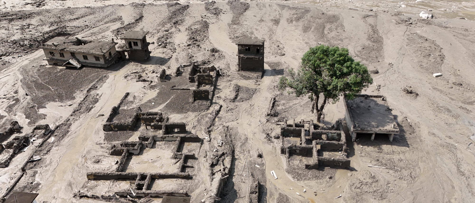 Luftaufnahme einer von Schlamm und Wasser überfluteten Landschaft. Mehrere Gebäude sind teilweise zerstört und von Schlamm umgeben. Ein einzelner grüner Baum steht neben den Ruinen. Die Umgebung zeigt Spuren von Erosion und Überschwemmung. Luftaufnahme einer von Schlamm und Wasser überfluteten Landschaft. Mehrere Gebäude sind teilweise zerstört und von Schlamm umgeben. Ein einzelner grüner Baum steht neben den Ruinen. Die Umgebung zeigt Spuren von Erosion und Überschwemmung.