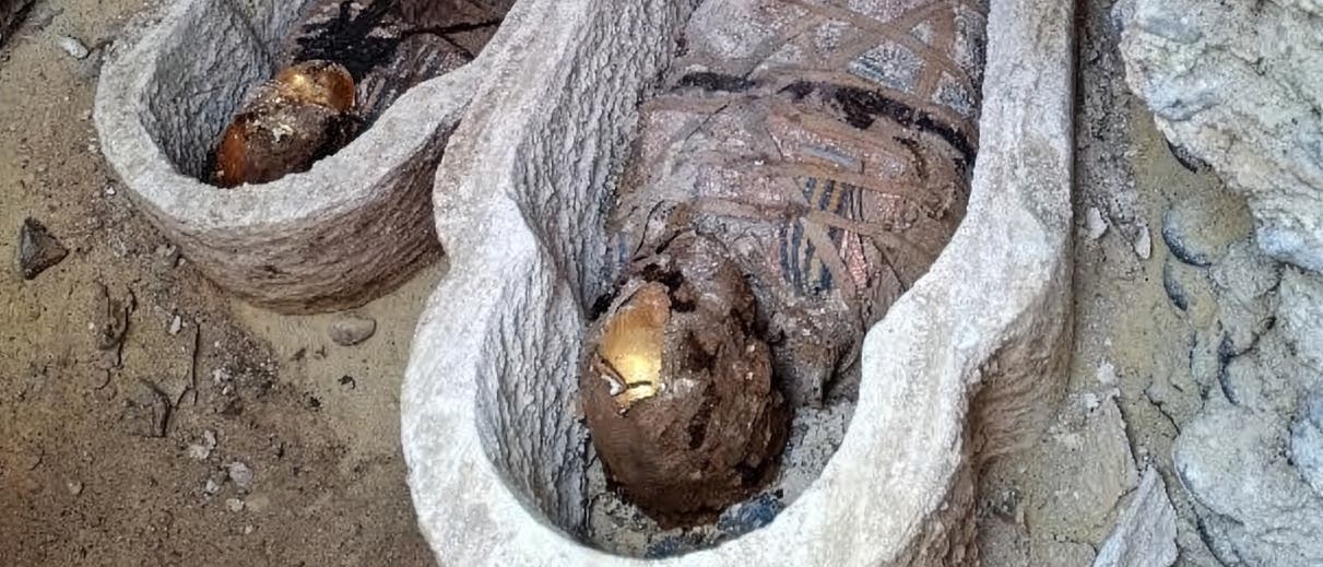 Zwei antike Mumien in steinernen Sarkophagen, die in einem arch&auml;ologischen Grabungsort entdeckt wurden. Die Mumien sind in Leinenbinden gewickelt, und ihre Gesichter sind mit goldenen Masken bedeckt. Die Umgebung zeigt sandigen Boden und Steinw&auml;nde, die auf eine historische St&auml;tte hinweisen.