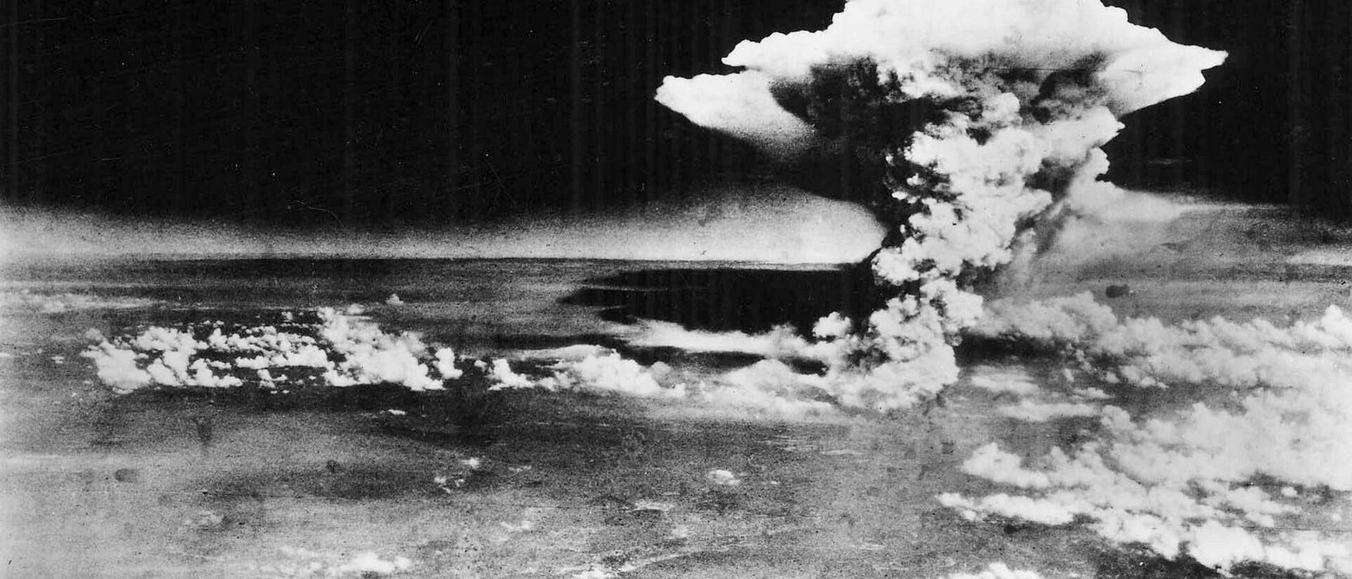 Eine Schwarz-Weiß-Fotografie zeigt eine große Pilzwolke, die in den Himmel aufsteigt, mit einer darunter liegenden Landschaft. Die Wolke besteht aus dichtem Rauch und Dampf, der sich nach oben ausbreitet. Die Szene vermittelt den Eindruck einer massiven Explosion. Im Vordergrund sind Teile einer Stadt und Küstenlinie zu erkennen.