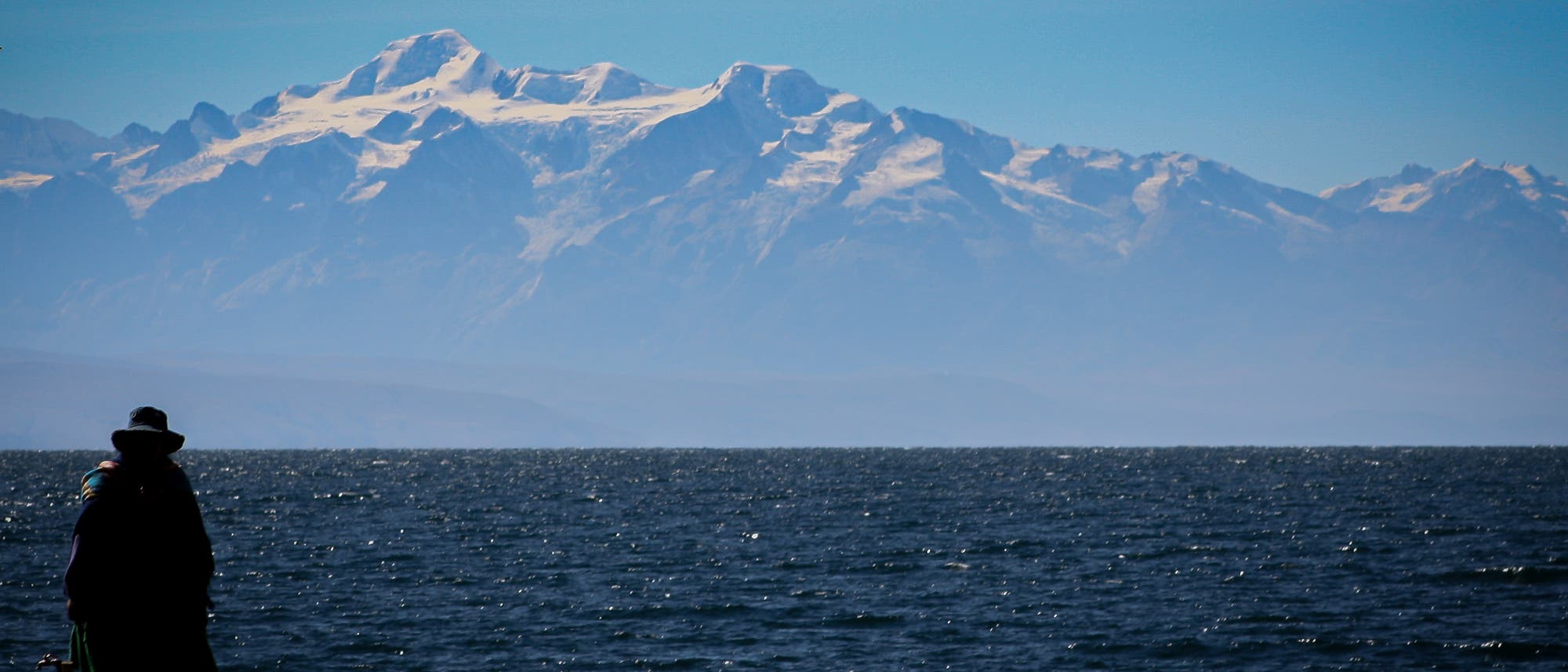 Eine Person steht am Ufer eines Sees, mit Blick auf eine beeindruckende Bergkette im Hintergrund. Die schneebedeckten Gipfel heben sich gegen den klaren blauen Himmel ab. Der See im Vordergrund reflektiert das Sonnenlicht, während die Silhouette der Person eine ruhige, nachdenkliche Stimmung vermittelt. Die Landschaft wirkt majestätisch und friedlich. Eine Person steht am Ufer eines Sees, mit Blick auf eine beeindruckende Bergkette im Hintergrund. Die schneebedeckten Gipfel heben sich gegen den klaren blauen Himmel ab. Der See im Vordergrund reflektiert das Sonnenlicht, während die Silhouette der Person eine ruhige, nachdenkliche Stimmung vermittelt. Die Landschaft wirkt majestätisch und friedlich.