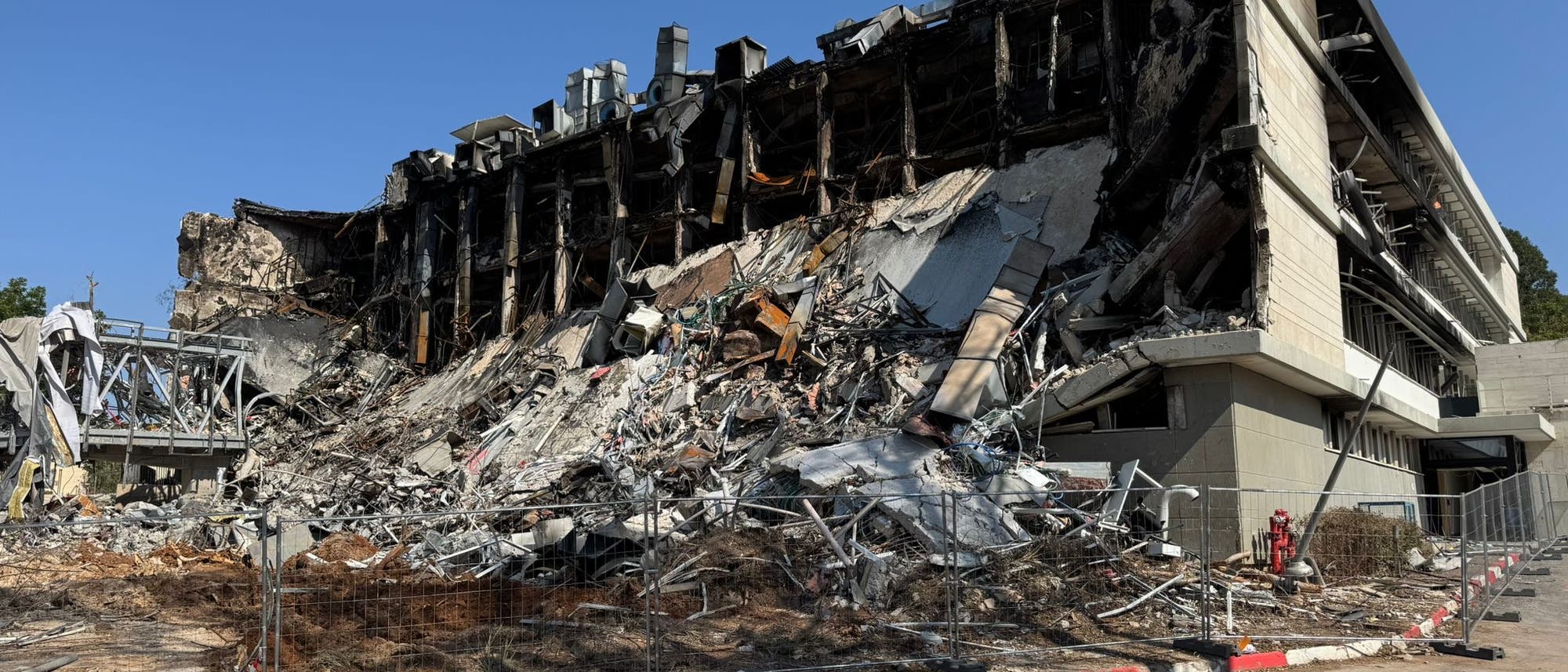 Ein teilweise eingestürztes Gebäude mit sichtbaren Trümmern und Stahlträgern. Die Fassade ist stark beschädigt, und es sind zahlreiche Schuttteile zu sehen. Ein Bauzaun umgibt die Ruine, und im Vordergrund ist eine Straße mit einem Zebrastreifen erkennbar. Der Himmel ist klar und blau.