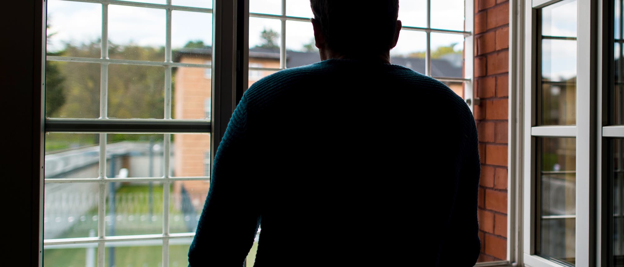 Eine Person steht mit dem Rücken zur Kamera und schaut aus einem großen, vergitterten Fenster in einen Hof mit Bäumen und einem Gebäude. Eine Person steht mit dem Rücken zur Kamera und schaut aus einem großen, vergitterten Fenster in einen Hof mit Bäumen und einem Gebäude.