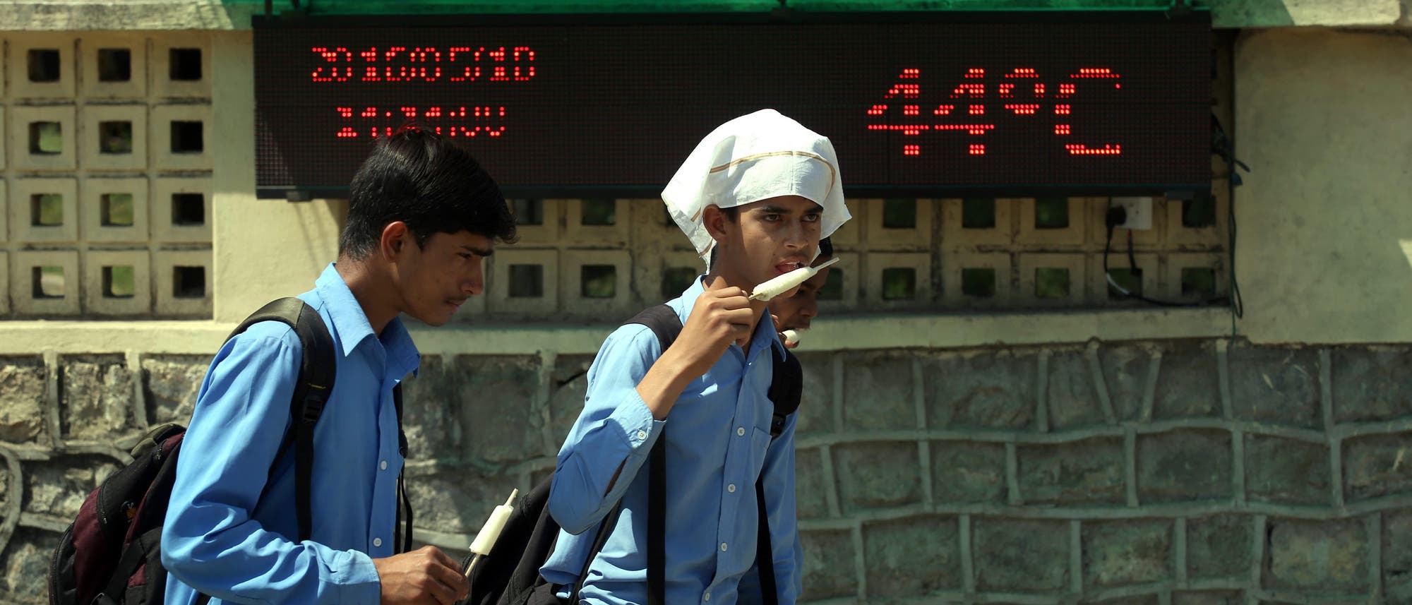 Indische Schüler essen Eis vor einer Anzeigetafel, die eine Temperatur von 44 Grad anzeigt.