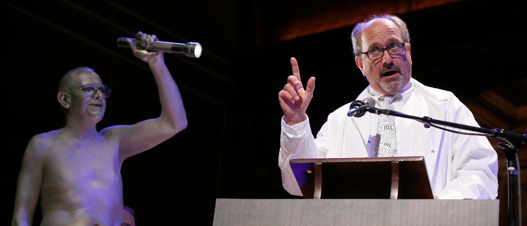 Ein Mann in einem wei&szlig;en Laborkittel steht an einem Rednerpult und spricht in ein Mikrofon. Neben ihm steht eine Person, die silbern bemalt ist und eine Taschenlampe hochh&auml;lt. Auf dem Rednerpult ist ein gro&szlig;es Schild mit der Aufschrift &raquo;Ig&laquo; zu sehen. Die Szene spielte sich bei der Ig-Nobelpreisverleihung 2016 ab.