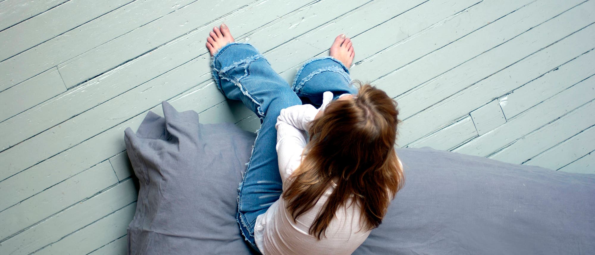 Eine Jugendliche mit langen, glatten, rotblonden Haaren sitzt auf einer grau bezogenen Matratze, die auf einem hellgrau gestrichenen Dielenboden liegt. Sie trägt eine Bluejeans und ein weißes Hemd und hat das Gesicht in den Händen vergraben. Die Perspektive ist so gewählt, dass man die triste Szene von oben, aus der Luft sieht. 