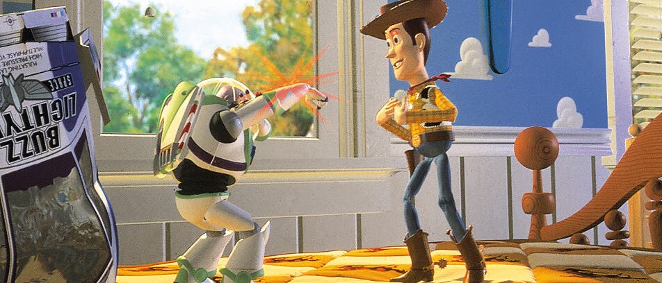 Eine animierte Szene zeigt zwei Spielzeugfiguren in einem Kinderzimmer. Links steht eine Figur in einem Raumanzug, die mit einem ausgestreckten Arm einen roten Laserstrahl abfeuert. Rechts steht eine Cowboy-Figur mit einem Hut, die lächelnd zuschaut. Im Hintergrund ist ein Fenster mit Blick auf Bäume und ein blauer Himmel mit Wolkenmuster an der Wand zu sehen. Auf dem Bett liegt eine bunte Decke mit Cowboy-Motiven. Links im Bild ist eine Verpackung mit der Aufschrift "Buzz Lightyear" zu erkennen.