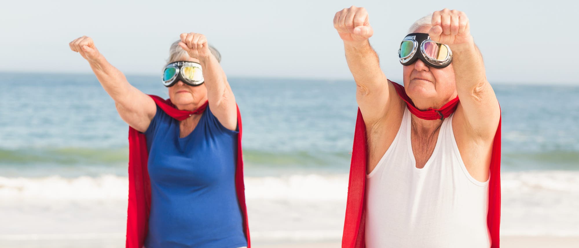 Zwei ältere Personen stehen am Strand und posieren mit ausgestreckten Armen, als ob sie fliegen würden. Sie tragen rote Umhänge und Schutzbrillen, was an Superhelden erinnert. Im Hintergrund ist das Meer zu sehen.