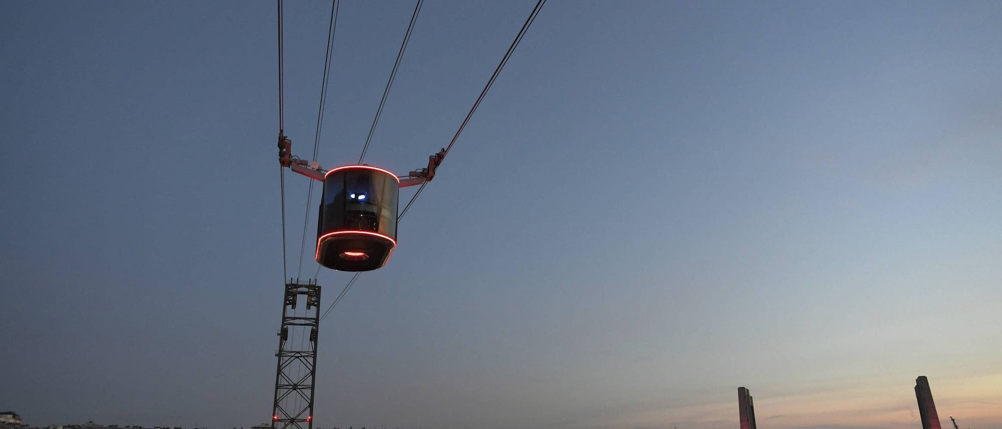 Seilbahn über einer Stadtlandschaft bei Dämmerung. Eine beleuchtete Kabine schwebt an Seilen, während im Hintergrund eine beleuchtete Brücke und Gebäude zu sehen sind. Der Himmel ist klar mit einem sichtbaren Mond. Seilbahn über einer Stadtlandschaft bei Dämmerung. Eine beleuchtete Kabine schwebt an Seilen, während im Hintergrund eine beleuchtete Brücke und Gebäude zu sehen sind. Der Himmel ist klar mit einem sichtbaren Mond.