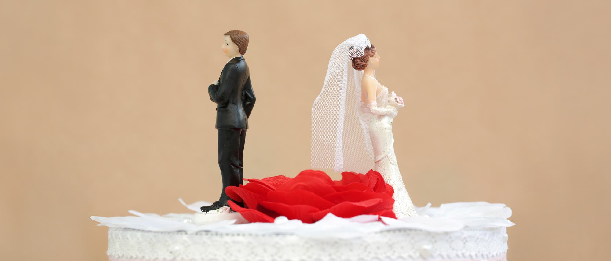 Auf einer Hochzeitstorte stehen zwei Plastikfiguren von Braut und Bräutigam, jedoch mit Abstand und einander den Rücken zugewandt, dazwischen eine rote Rose.