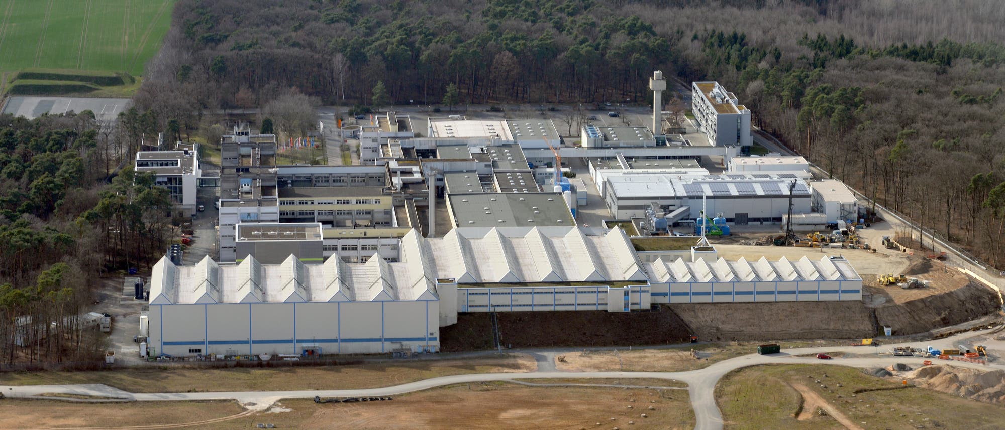 Luftaufnahme eines gro&szlig;en Industriekomplexes, umgeben von Wald und Feldern. Die Geb&auml;ude haben wei&szlig;e D&auml;cher und sind in einem rechteckigen Muster angeordnet. Im Vordergrund sind unbebaute Fl&auml;chen und ein kurviger Weg zu sehen. Im Hintergrund erstrecken sich dichte W&auml;lder.