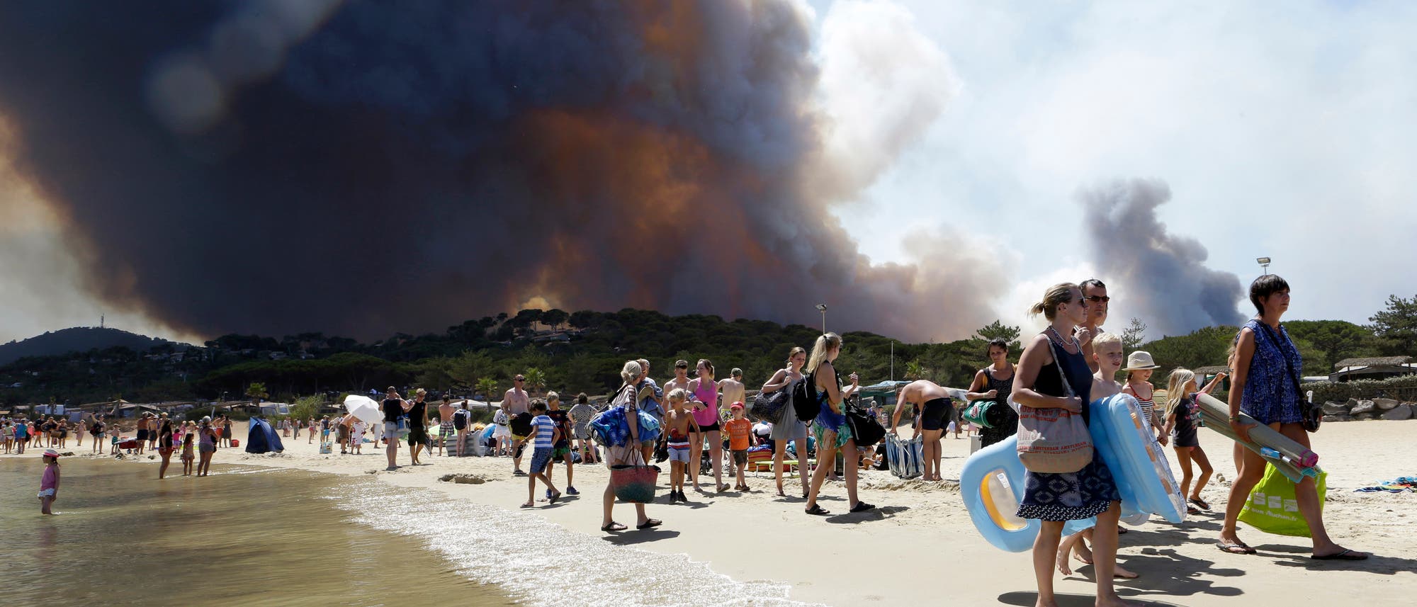 Ein Strand wird evakuiert, während hinter den Dünen im Hintergrund bereits dunkler Rauch eines Waldbrands aufsteigt.