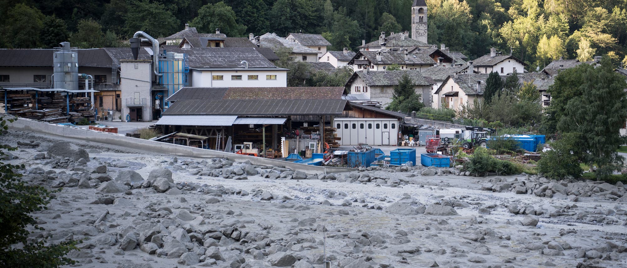 Eine kleine, von Bäumen umgebene Ortschaft mit traditionellen Steinhäusern und einer Kirche im Hintergrund. Im Vordergrund sind Industriegebäude und Container zu sehen. Der Boden ist mit Geröll und Schlamm bedeckt, was auf einen Erdrutsch oder eine Überschwemmung hindeutet. Eine kleine, von Bäumen umgebene Ortschaft mit traditionellen Steinhäusern und einer Kirche im Hintergrund. Im Vordergrund sind Industriegebäude und Container zu sehen. Der Boden ist mit Geröll und Schlamm bedeckt, was auf einen Erdrutsch oder eine Überschwemmung hindeutet.