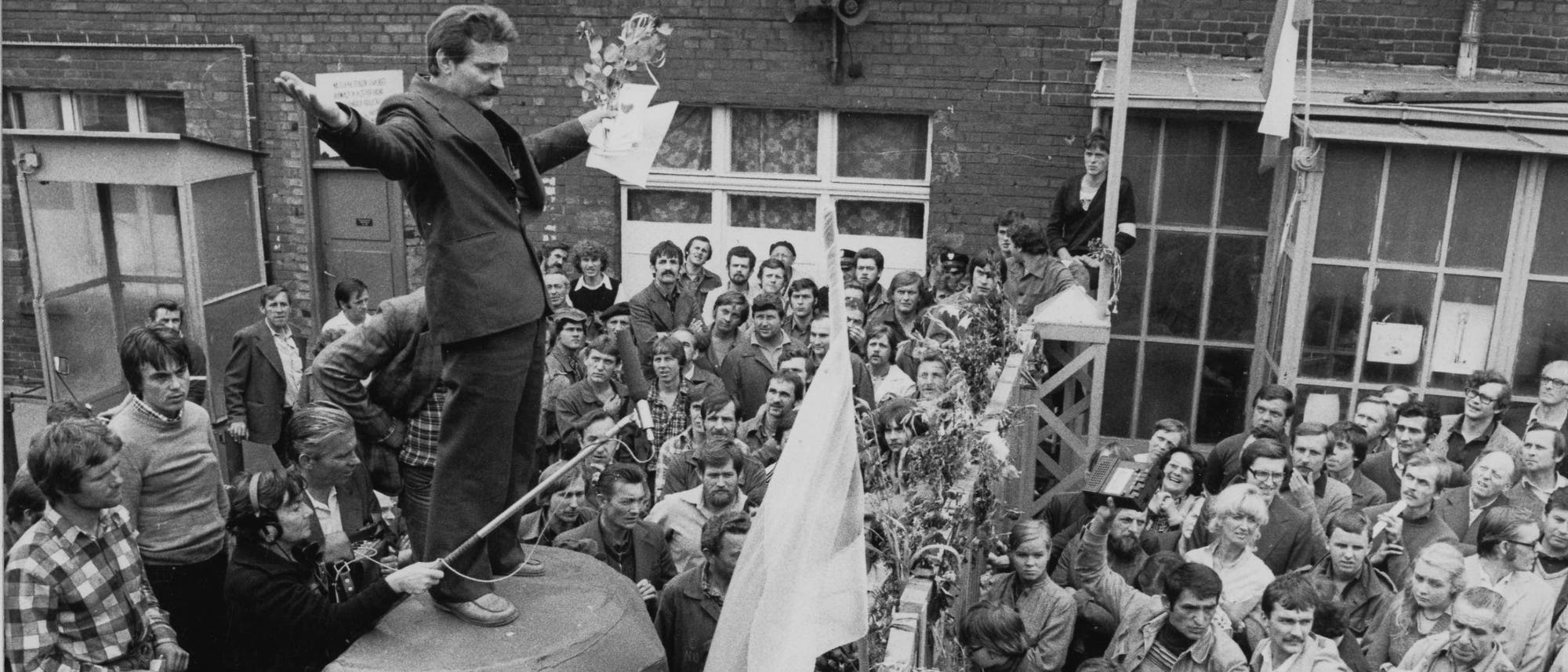 In der Schwarz-weiß-Aufnahme steht der junge Lech Wałęsa auf einem kleinen Podium inmitten von Männern und Frauen. Er trägt ein dunkles Sakko und hat beide Arme zur Seite ausgestreckt.