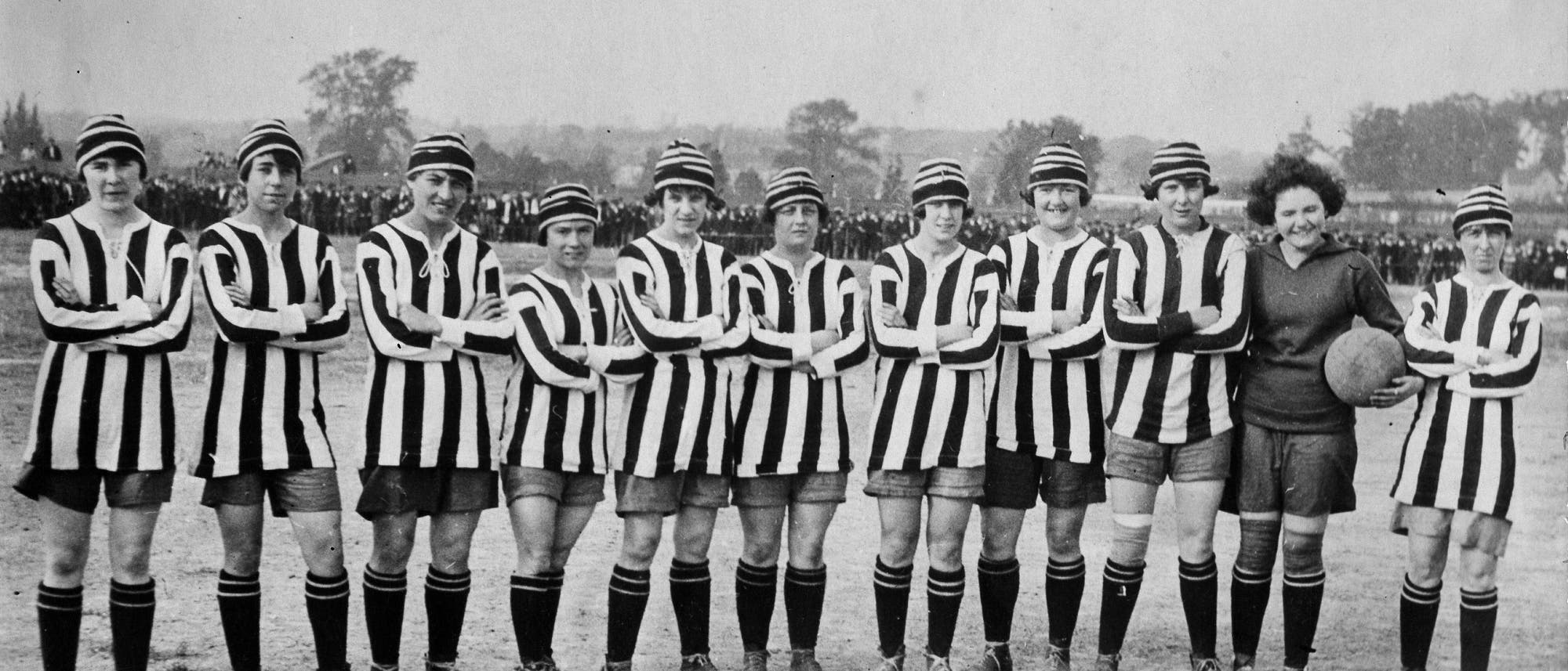 Eine Gruppe von Frauen in gestreiften Fußballtrikots und Mützen steht in einer Reihe auf einem Fußballfeld. Sie haben die Arme verschränkt und lächeln in die Kamera. Eine Frau hält einen Fußball. Im Hintergrund sind Zuschauer zu sehen. Die Szene vermittelt den historischen Kontext eines Frauenfußballteams. Eine Gruppe von Frauen in gestreiften Fußballtrikots und Mützen steht in einer Reihe auf einem Fußballfeld. Sie haben die Arme verschränkt und lächeln in die Kamera. Eine Frau hält einen Fußball. Im Hintergrund sind Zuschauer zu sehen. Die Szene vermittelt den historischen Kontext eines Frauenfußballteams.
