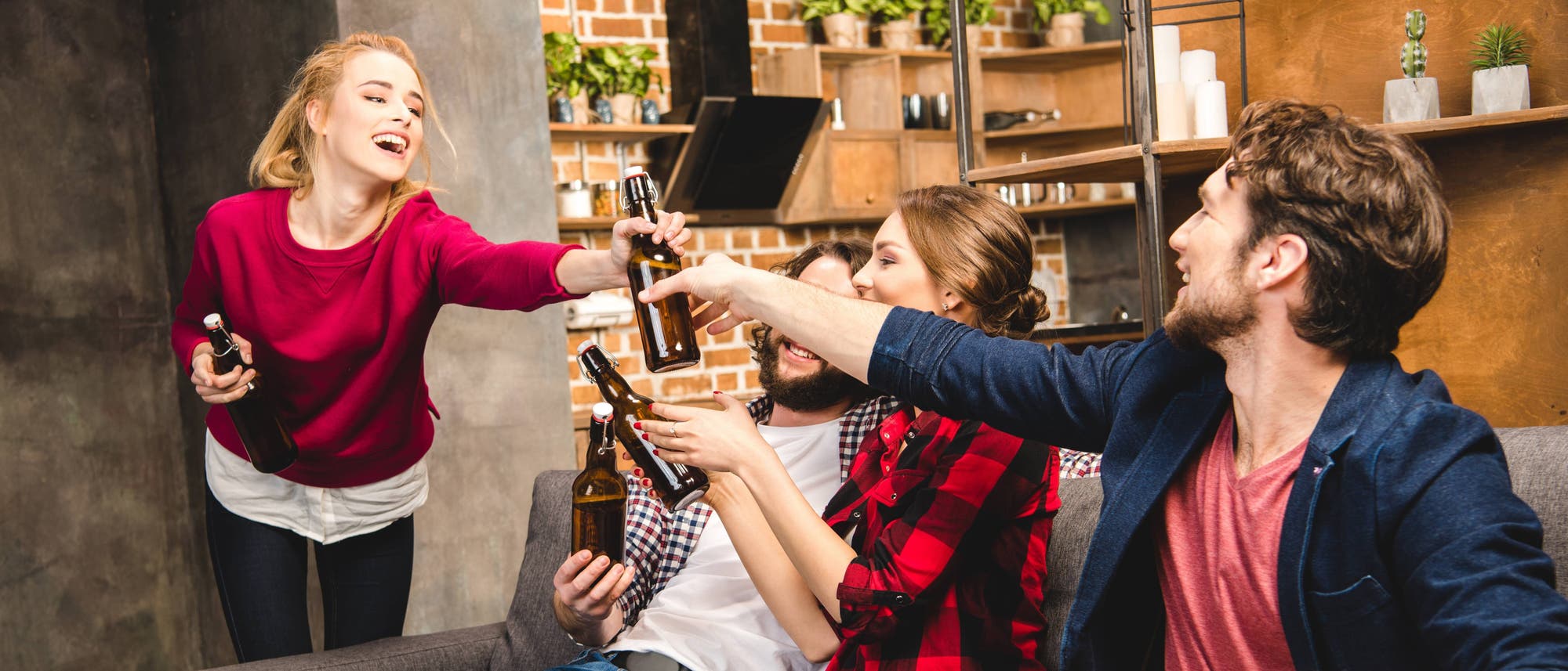 Eine Gruppe von vier jungen Erwachsenen in einem gemütlichen Wohnzimmer mit Backsteinwänden und Regalen voller Pflanzen. Sie lachen und stoßen mit Bierflaschen an. Eine Frau im roten Pullover, die als einzige steht, reicht einem Mann auf einer Couch eine Bierflasche, während die anderen zwei Personen ebenfalls sitzen und Flaschen in der Hand halten. Die Szene vermittelt eine fröhliche und gesellige Atmosphäre. Eine Gruppe von vier jungen Erwachsenen in einem gemütlichen Wohnzimmer mit Backsteinwänden und Regalen voller Pflanzen. Sie lachen und stoßen mit Bierflaschen an. Eine Frau im roten Pullover, die als einzige steht, reicht einem Mann auf einer Couch eine Bierflasche, während die anderen zwei Personen ebenfalls sitzen und Flaschen in der Hand halten. Die Szene vermittelt eine fröhliche und gesellige Atmosphäre.