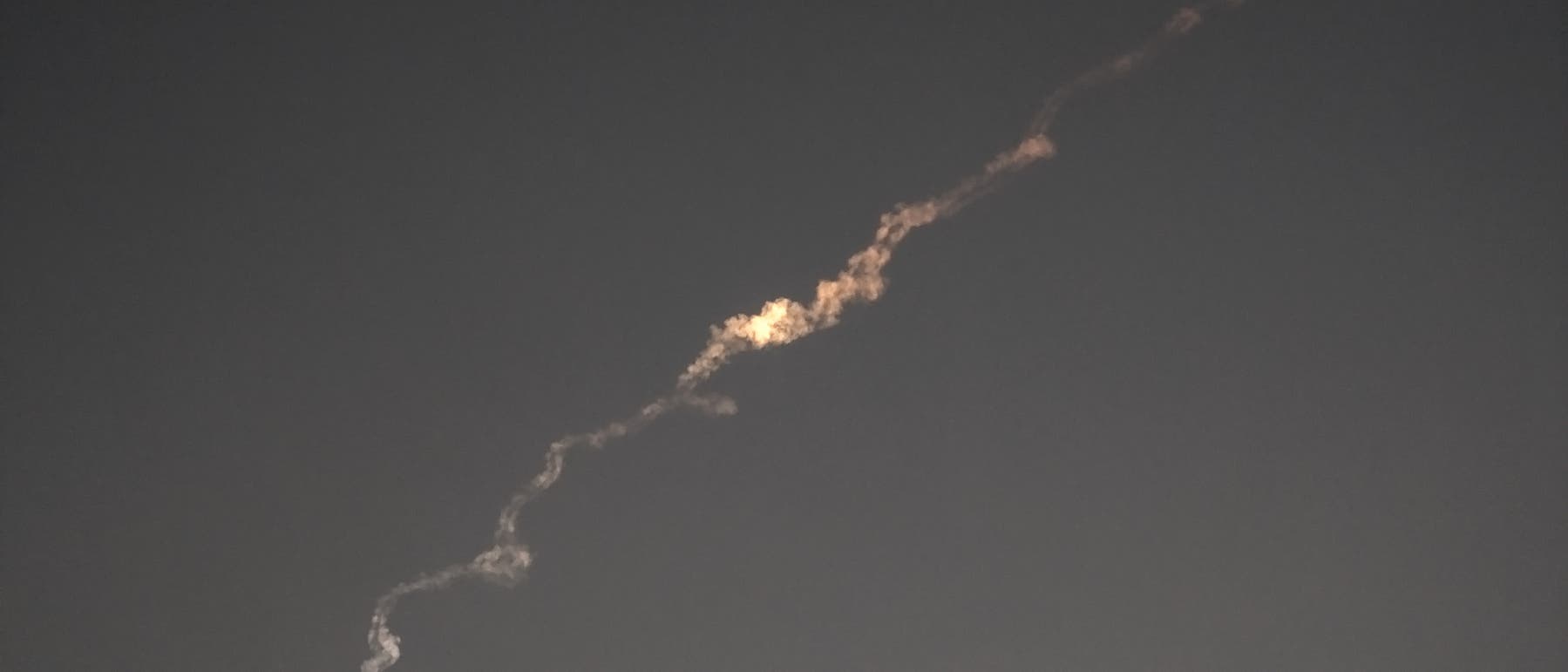 Ein heller, gewundener Kondensstreifen zieht sich durch den dunklen Himmel, m&ouml;glicherweise von einem Raketenstart oder Flugzeug. Der Streifen leuchtet in der Mitte intensiver, was auf eine Lichtquelle oder Reflexion hindeutet.