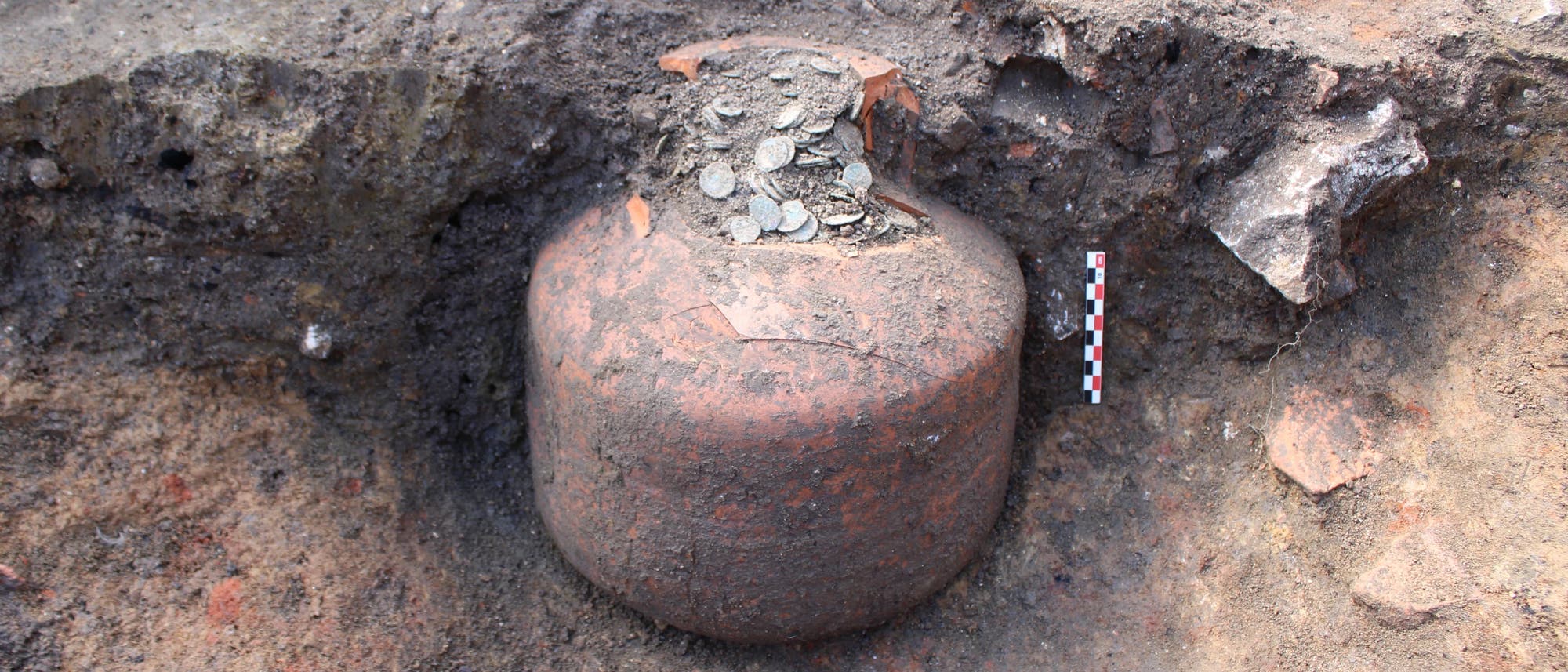Eine archäologische Ausgrabung zeigt einen großen, teilweise freigelegten Tonkrug, der mit alten Münzen gefüllt ist. Der Krug ist in der Erde eingebettet, umgeben von Erd- und Gesteinsschichten. Rechts neben dem Krug befindet sich eine Messlatte zur Größenbestimmung. Die Szene vermittelt den Eindruck einer historischen Entdeckung.
