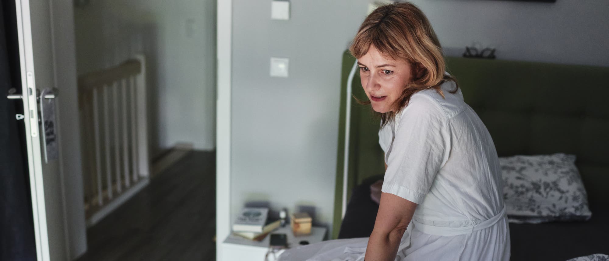 Eine Person in einem weißen Kleid sitzt auf einem Bett in einem Schlafzimmer. Der Raum ist in neutralen Tönen gehalten, mit einem Nachttisch, auf dem Bücher und kleine Gegenstände liegen. Eine offene Tür führt zu einem Flur. Die Person schaut nachdenklich zur Seite.