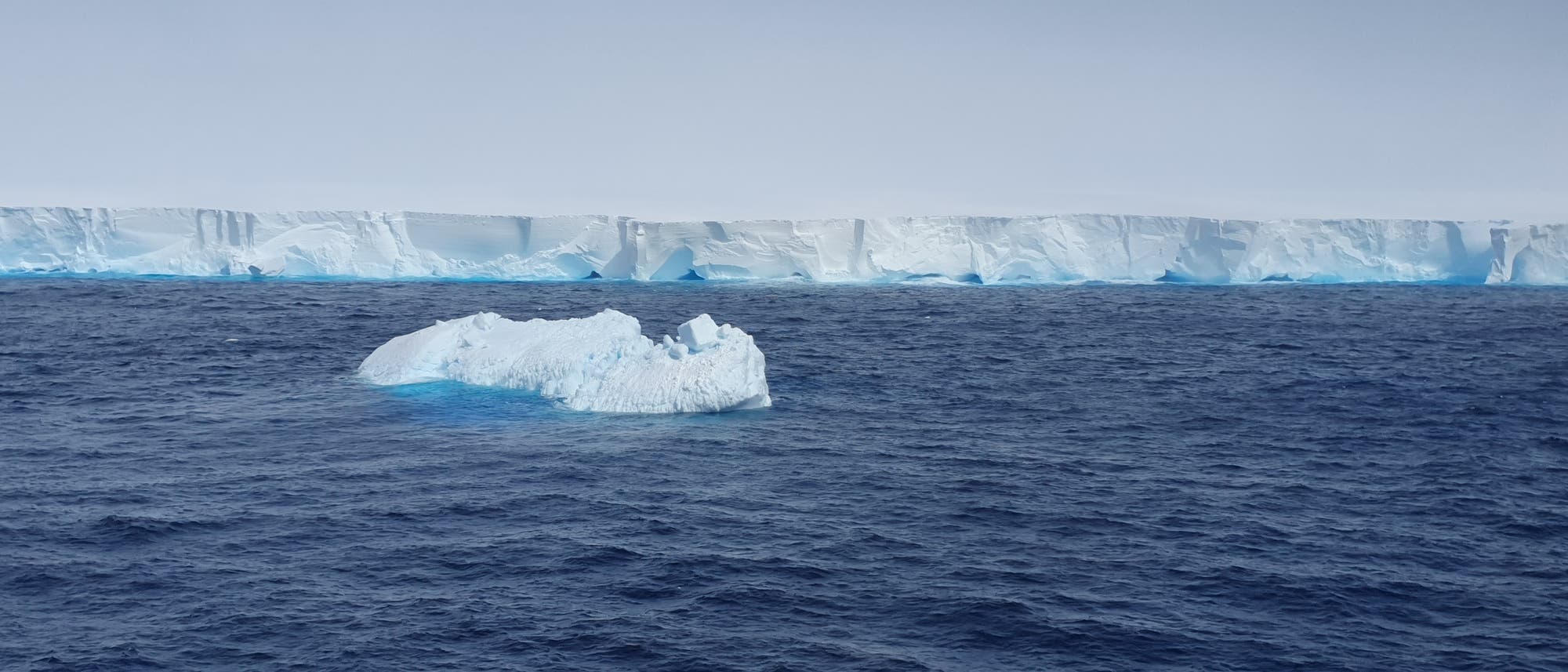 Eine weiße Eiswand ragt aus dem blauen Südpolarmeer, in dem ein viel kleinerer Eisberg zusätzlich schwimmt.