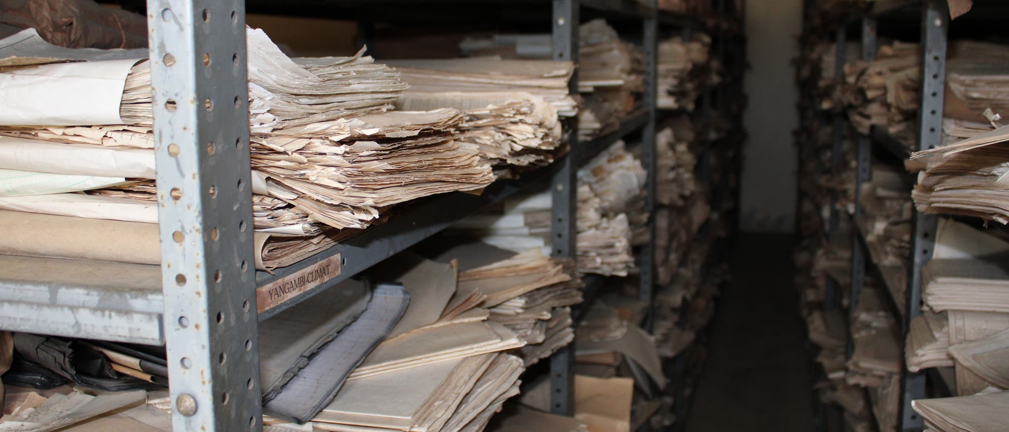 Regale voller alter Papierdokumente in einem Archivraum in der Demokratischen Republik Kongo. Die Dokumente sind gestapelt und teilweise vergilbt. Im Raum stehen mehrere Reihen von Metallregalen eng nebeneinander.