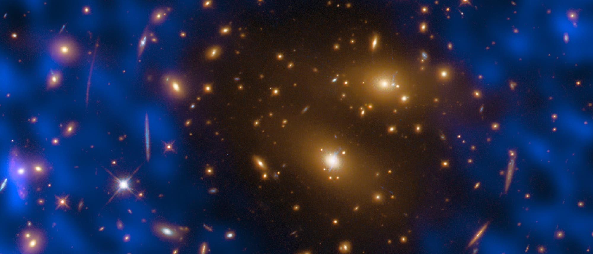 Ein astronomisches Bild zeigt einen Galaxienhaufen im Weltraum. Im Zentrum sind zahlreiche leuchtende, gelbliche Galaxien zu sehen, die von einem blauen, nebligen Hintergrund umgeben sind. Die blauen Bereiche deuten auf die Verteilung von Dunkler Materie hin. Das Bild veranschaulicht die Gravitationslinsenwirkung, bei der das Licht entfernter Galaxien durch die Masse des Haufens verzerrt wird. Dieses Ph&auml;nomen wird durch die verzerrten Lichtb&ouml;gen und Streifen um den Haufen herum sichtbar.