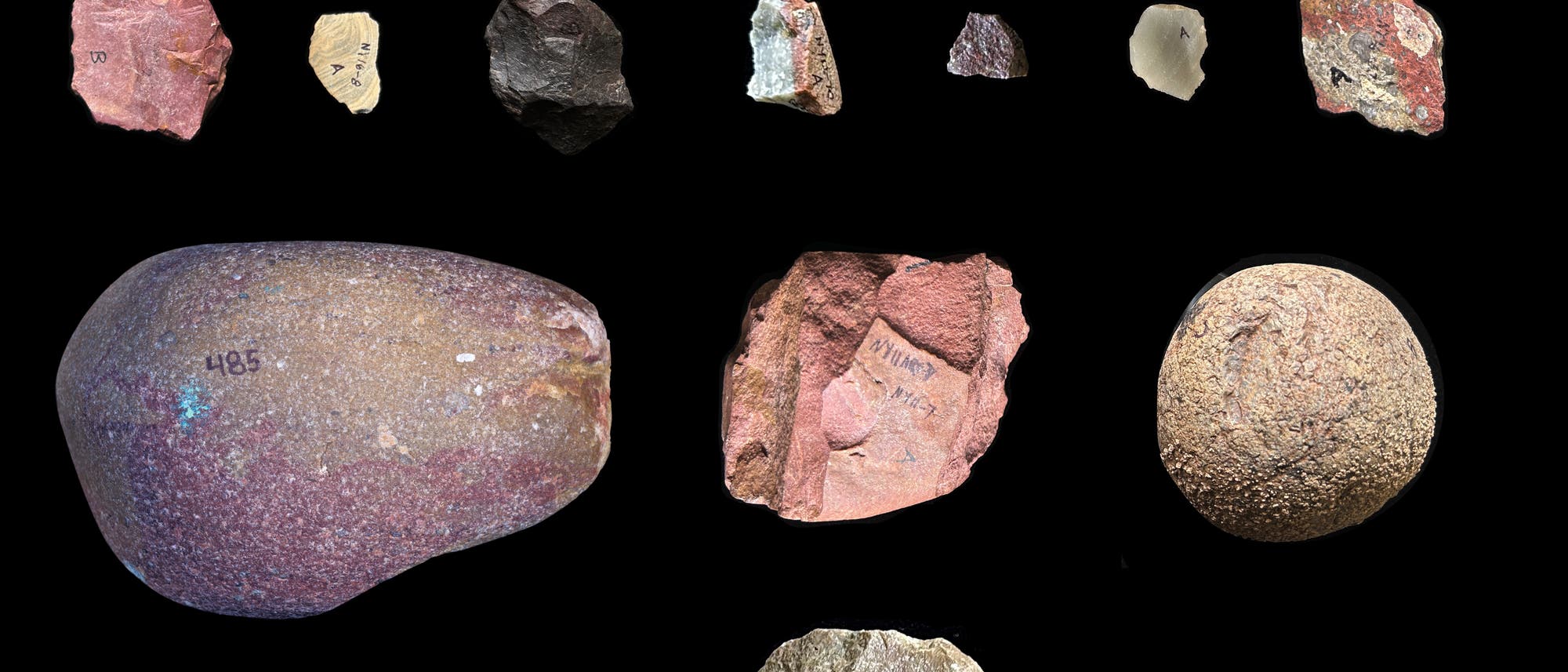 Eine Sammlung von 18 Steingeräten, die auf einem schwarzen Hintergrund angeordnet sind. Die Proben variieren in Form, Größe und Farbe, von rötlichen bis zu erdigen Tönen. Einige Steine zeigen glatte Oberflächen, während andere rau und unregelmäßig sind. In der unteren rechten Ecke befindet sich eine Maßstabsleiste von 2 cm. Eine Sammlung von 18 Steingeräten, die auf einem schwarzen Hintergrund angeordnet sind. Die Proben variieren in Form, Größe und Farbe, von rötlichen bis zu erdigen Tönen. Einige Steine zeigen glatte Oberflächen, während andere rau und unregelmäßig sind. In der unteren rechten Ecke befindet sich eine Maßstabsleiste von 2 cm.