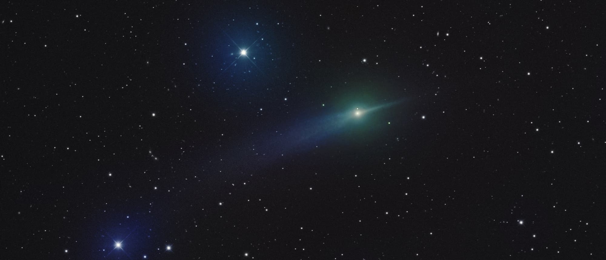 Ein astronomisches Bild zeigt einen Kometen mit einem leuchtend gr&uuml;nen Kopf und einem langen, bl&auml;ulichen Schweif, der sich durch ein Sternenfeld zieht. Zwei helle Sterne sind in der N&auml;he des Kometen sichtbar, einer links oben und der andere links unten. Der Hintergrund ist mit zahlreichen kleineren Sternen &uuml;bers&auml;t, die den Nachthimmel darstellen. Der Komet und die Sterne heben sich deutlich vom dunklen Hintergrund ab.