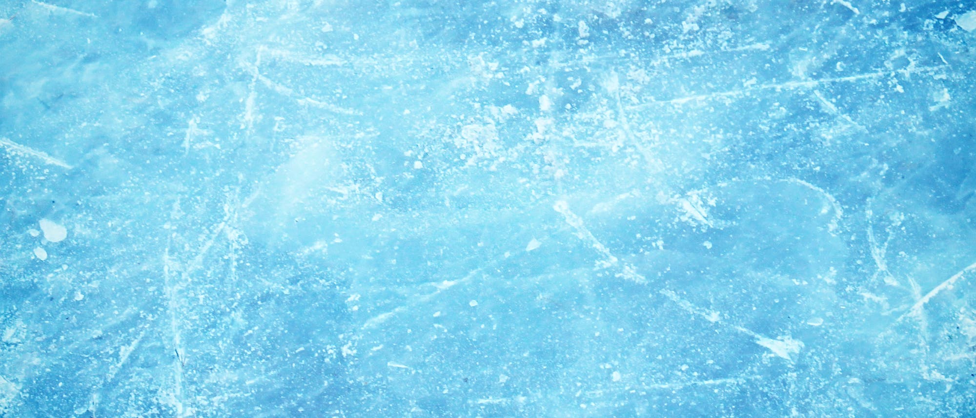 Eine Nahaufnahme einer eisigen Oberfläche mit einer hellblauen Tönung. Die Textur zeigt Kratzer und unregelmäßige Muster, die an gefrorenes Wasser erinnern. Die Oberfläche wirkt rau und uneben, was auf eine natürliche Eisbildung hinweist.