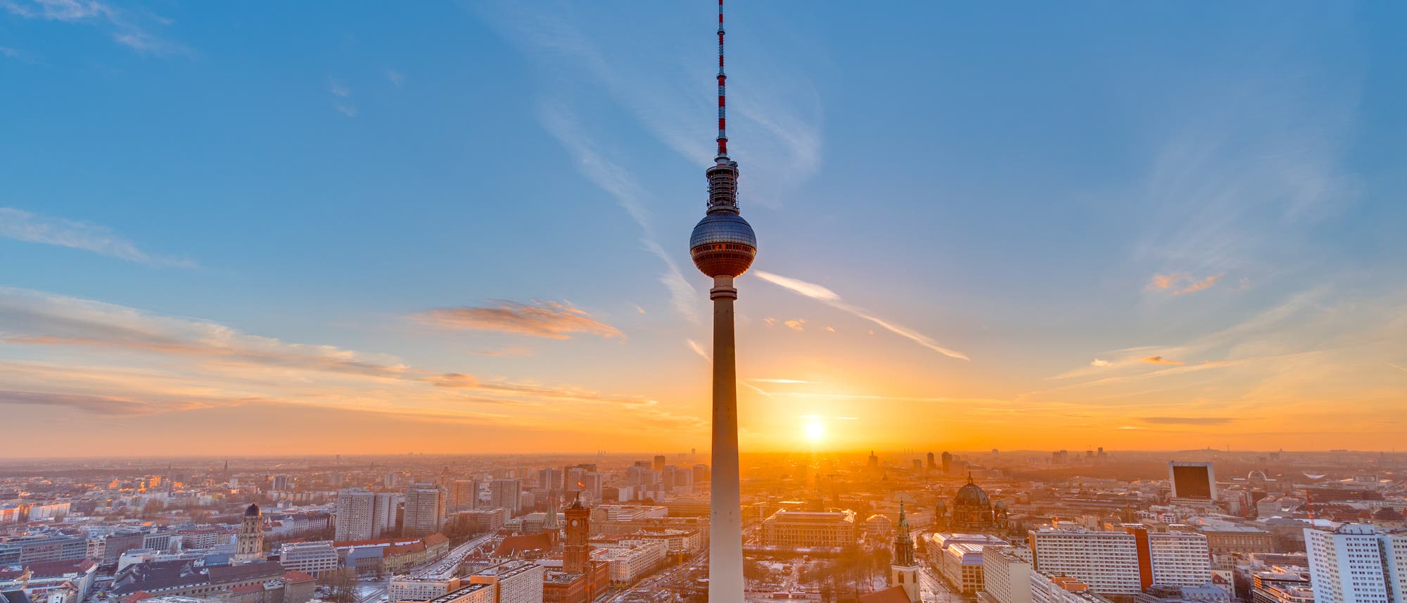 Stadtansicht von Berlin bei Sonnenuntergang, mit dem Fernsehturm im Vordergrund. Der Himmel ist klar mit vereinzelten Wolken, und die Sonne geht am Horizont unter, was die Gebäude in warmes Licht taucht. Die Stadtlandschaft zeigt eine Mischung aus modernen und historischen Gebäuden.