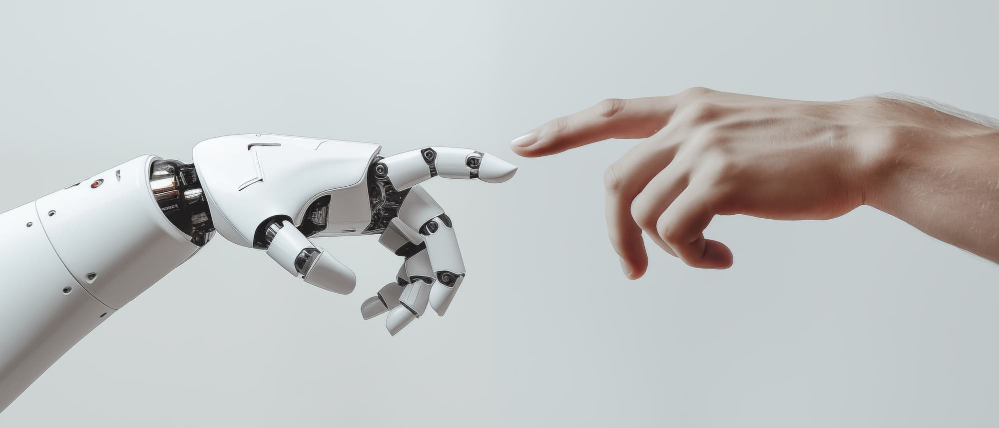 Eine menschliche Hand und eine Roboterhand nähern sich einander, wobei die Fingerspitzen fast in Berührung kommen. Die Szene erinnert an das berühmte Gemälde "Die Erschaffung Adams" und symbolisiert die Verbindung zwischen Mensch und Technologie. Der Hintergrund ist neutral und lenkt nicht vom Hauptmotiv ab.