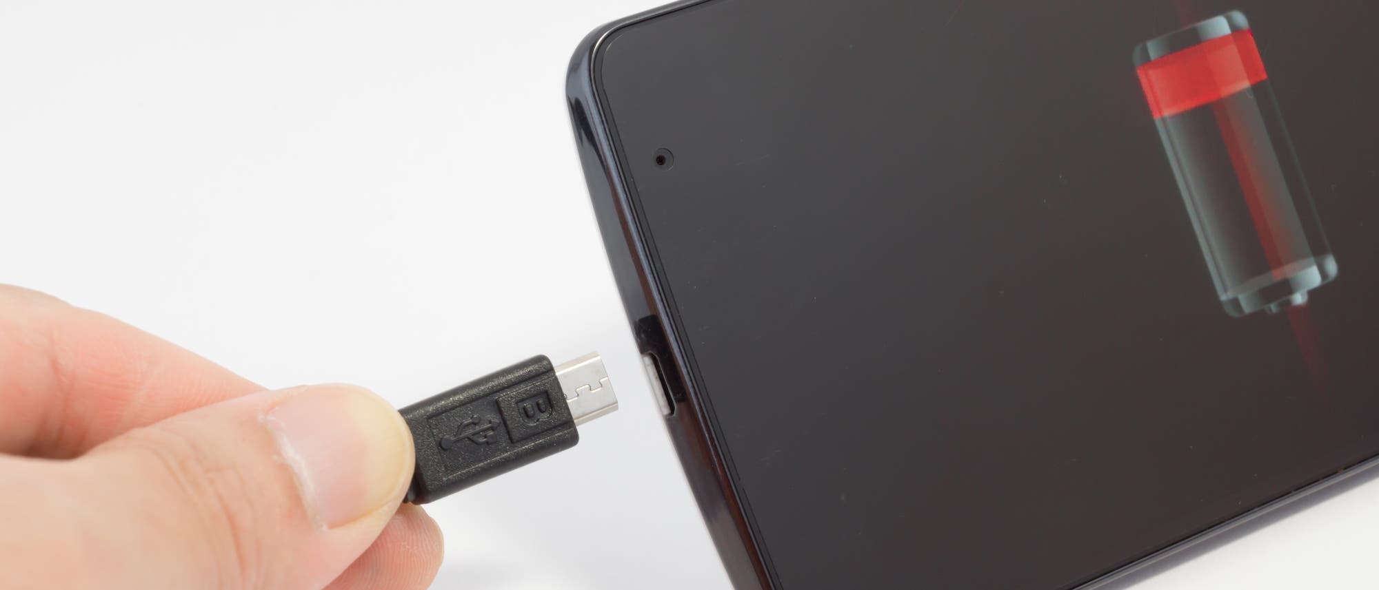 Ein Mobiltelefon mit roter Ladeanzeige, an das eine Hand gerade das Ladekabel anschließt. Ein Mobiltelefon mit roter Ladeanzeige, an das eine Hand gerade das Ladekabel anschließt.