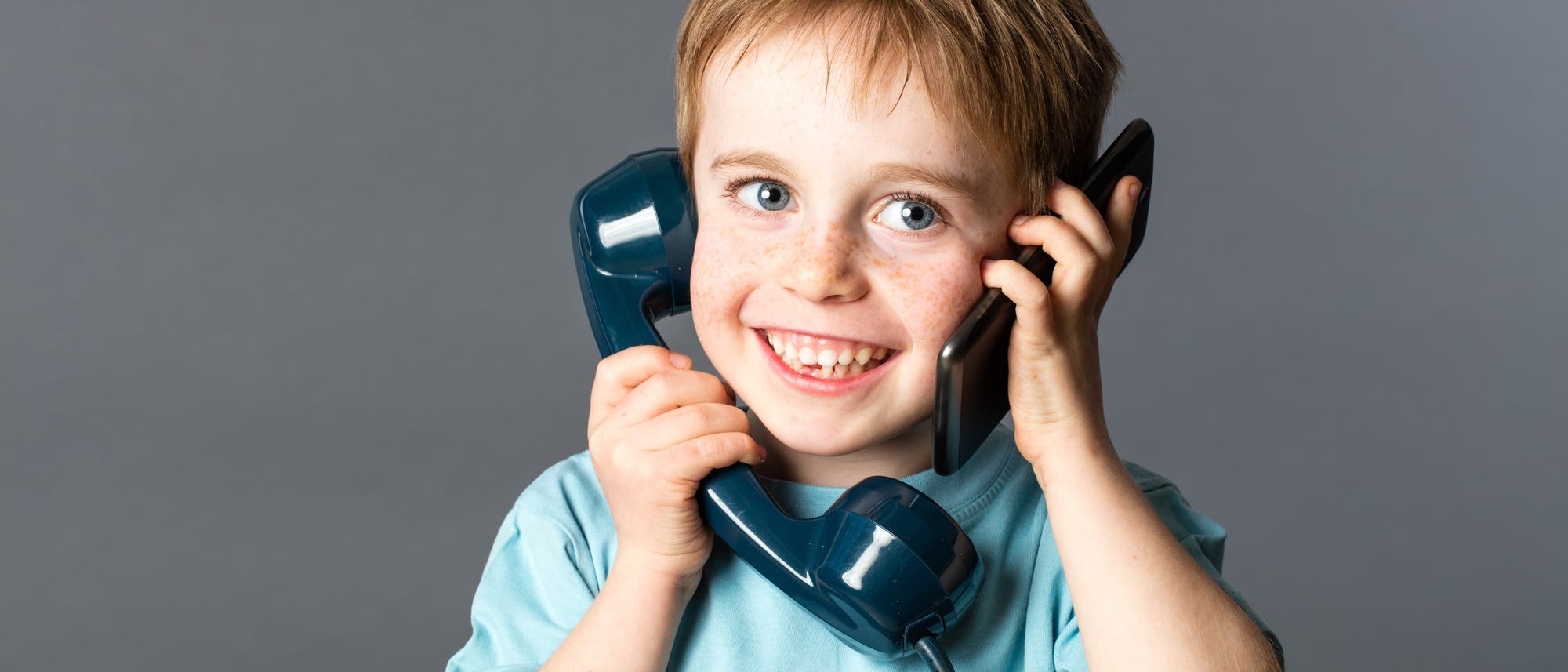Ein Kind lächelt fröhlich und hält zwei Telefone an die Ohren, eines in jeder Hand. Das linke Telefon ist ein klassischer, dunkelblauer Hörer, während das rechte ein modernes Smartphone ist. Der Hintergrund ist neutral grau, was den Fokus auf das Kind und die Telefone lenkt. Ein Kind lächelt fröhlich und hält zwei Telefone an die Ohren, eines in jeder Hand. Das linke Telefon ist ein klassischer, dunkelblauer Hörer, während das rechte ein modernes Smartphone ist. Der Hintergrund ist neutral grau, was den Fokus auf das Kind und die Telefone lenkt.