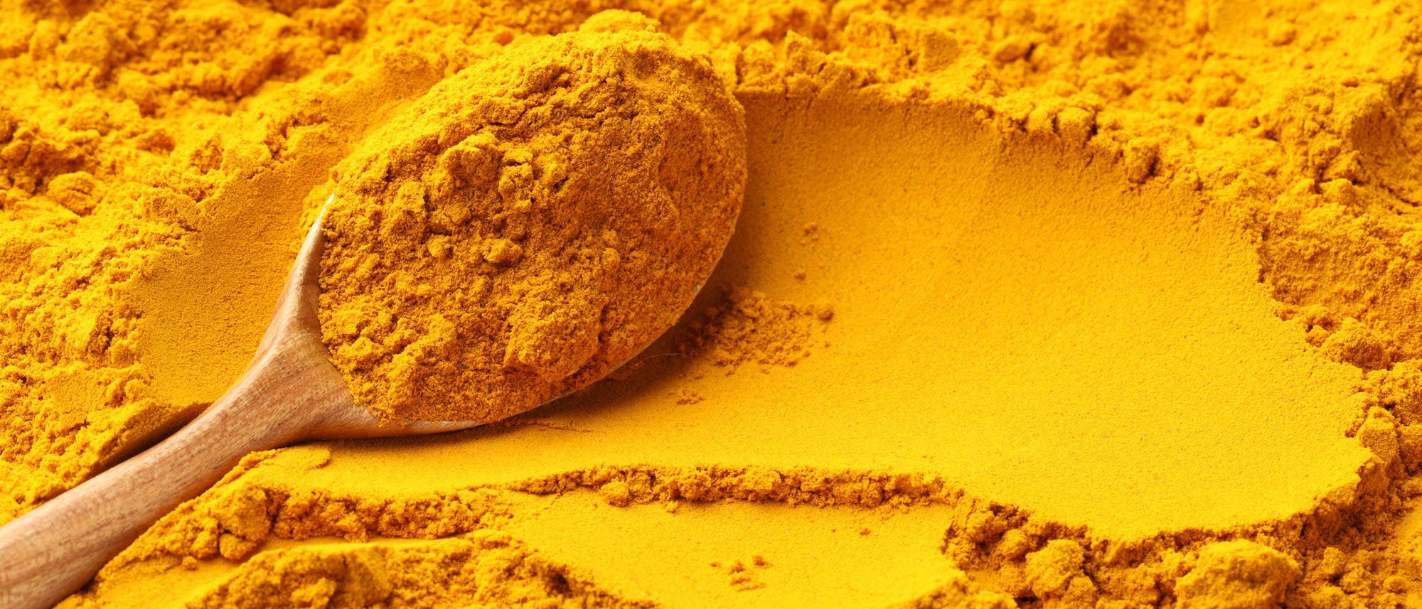 Ein Holzl&ouml;ffel liegt auf einer Oberfl&auml;che aus leuchtend gelbem Pulver, das sich um den L&ouml;ffel herum verteilt. Das Bild zeigt eine Nahaufnahme von Kurkumapulver, das f&uuml;r seine intensive Farbe und Verwendung in der K&uuml;che bekannt ist.