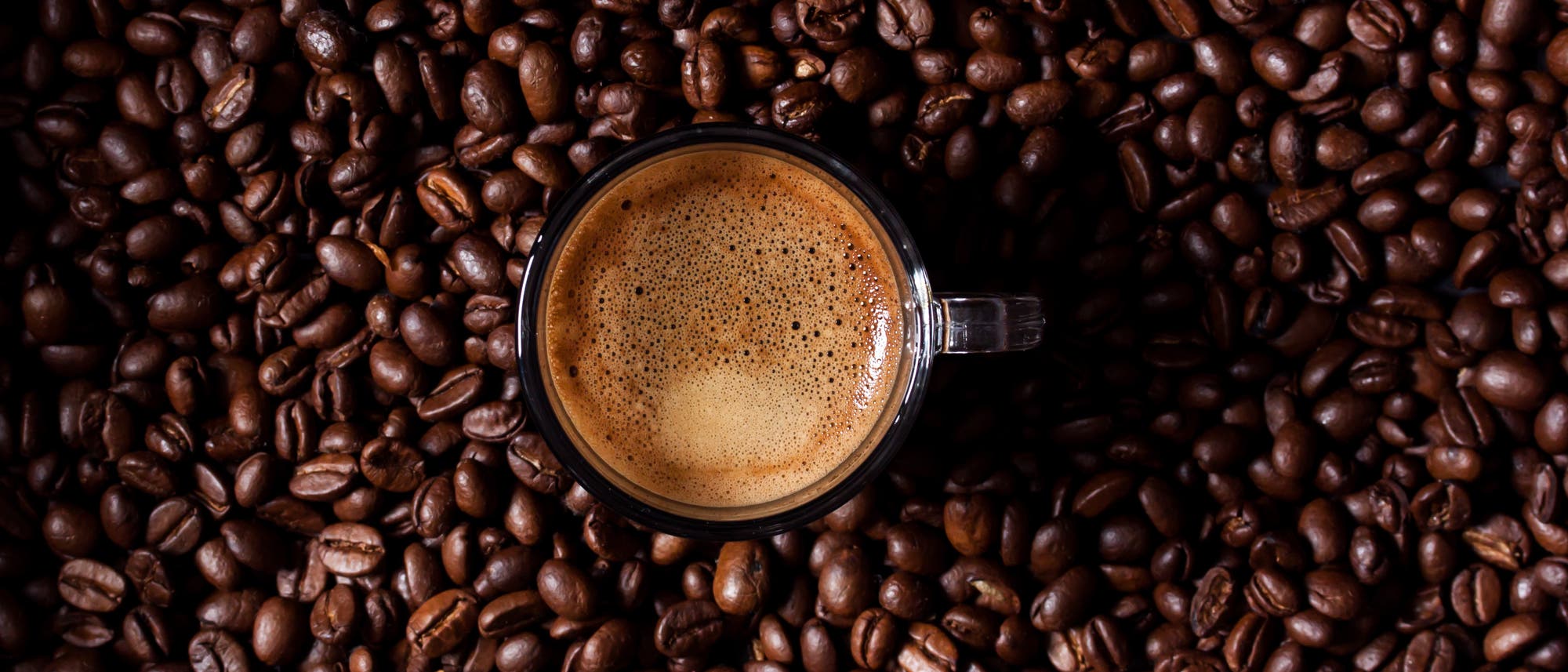 Eine Tasse Espresso mit einer cremigen Schaumkrone steht auf einer Oberfläche, die vollständig mit gerösteten Kaffeebohnen bedeckt ist.