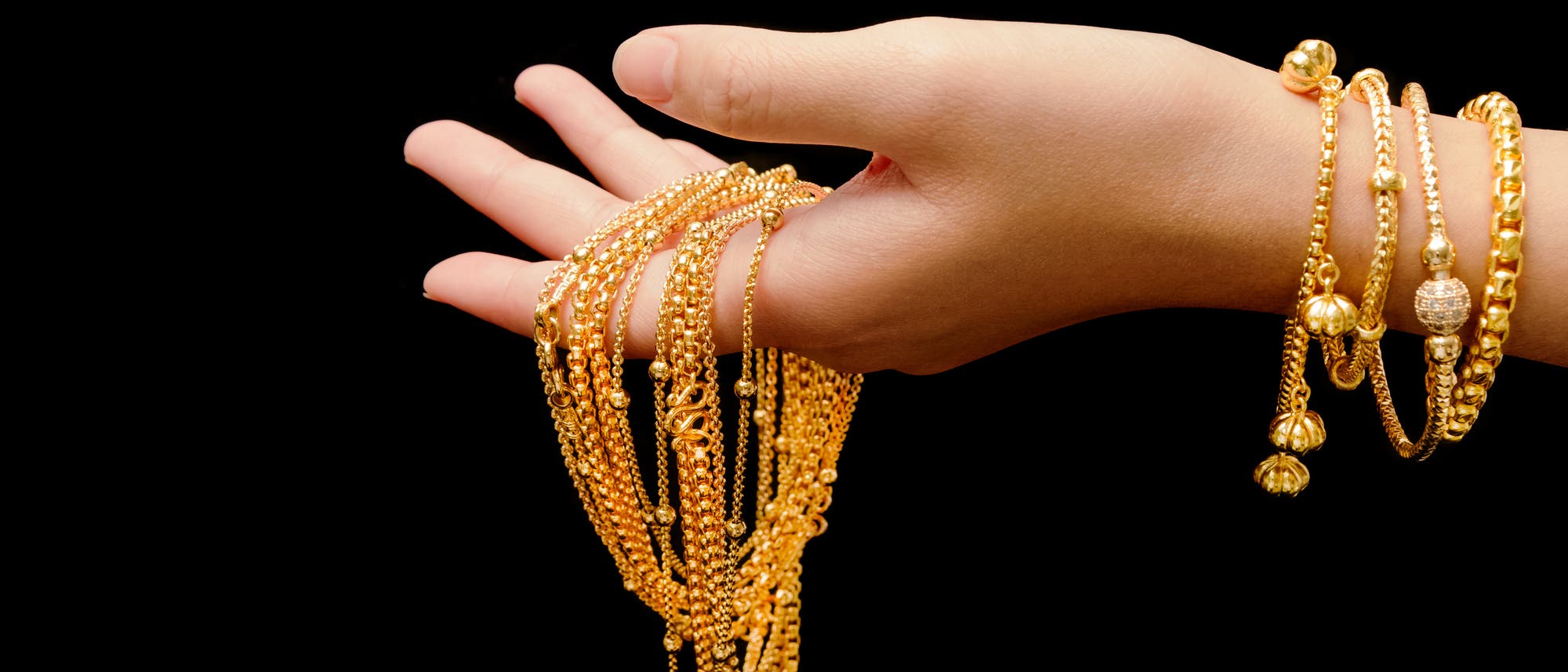 Eine Hand mit goldenen Armreifen und Ketten vor schwarzem Hintergrund.