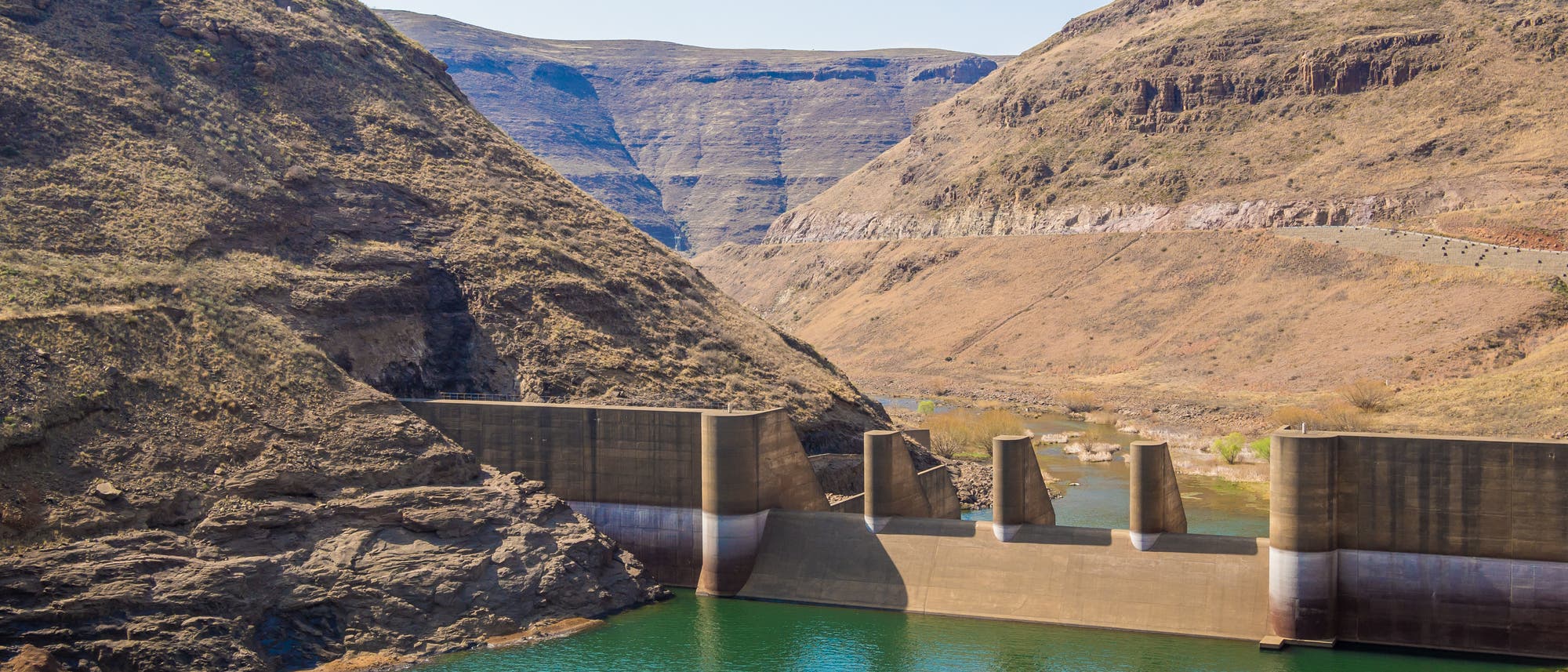 Ansicht des Wasserkraftwerks in Lesotho zwischen kargen Bergen, das Wasser ist grün-blau.